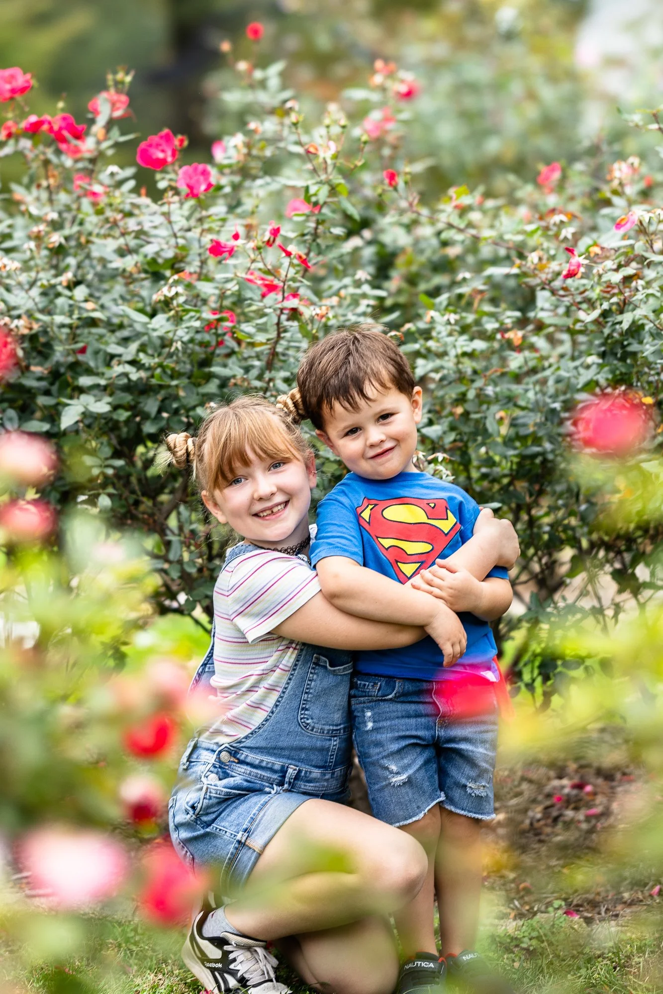 Two children, a girl and a boy, smiling and hugging each other among pink roses in a garden.
