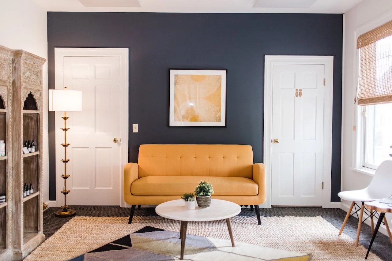 Living room with a dark accent wall, a yellow sofa, a white coffee table with potted plants, two white doors, a framed artwork, a tall lamp, a wooden shelf on the left, and a window with a bamboo shade on the right.