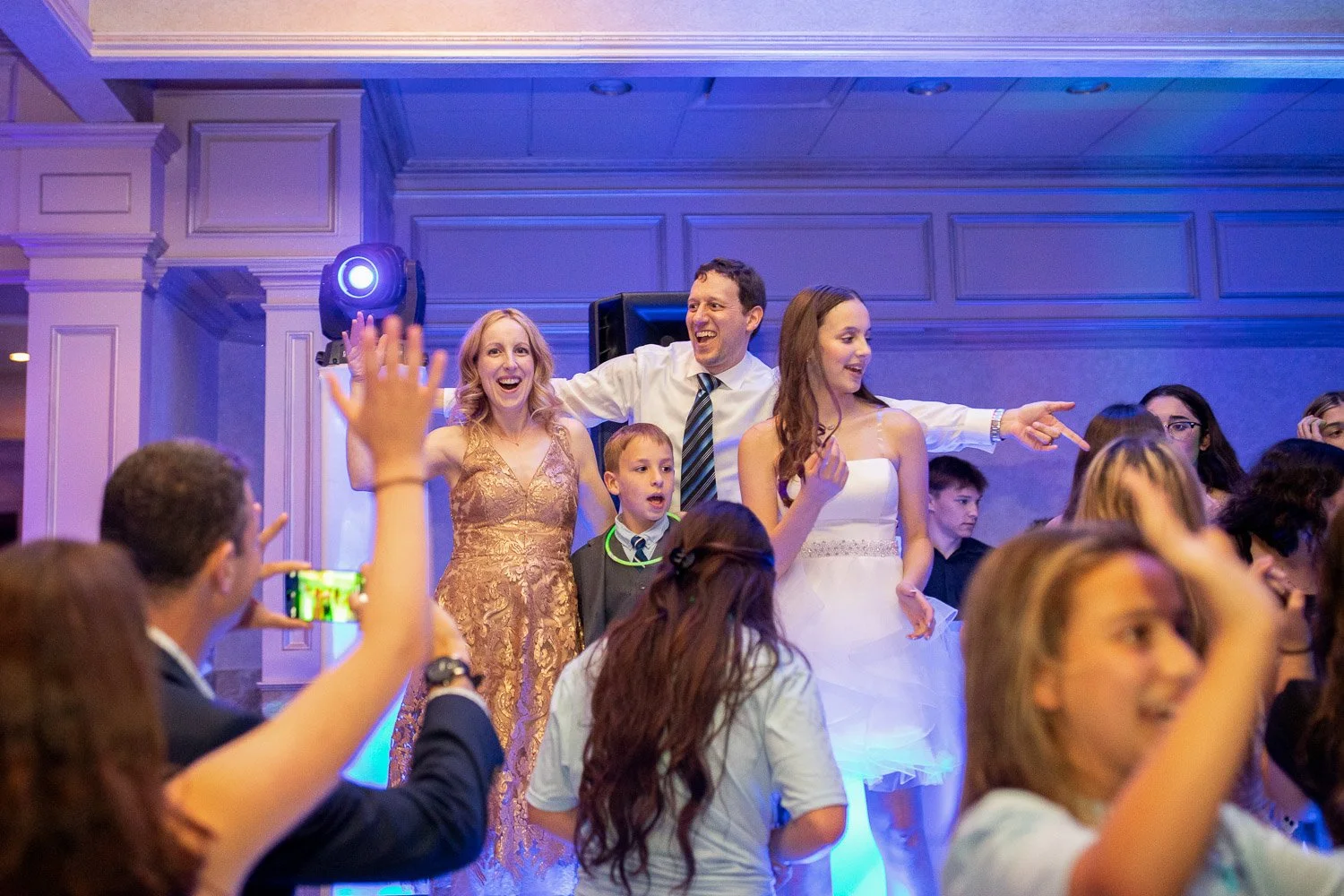 People dancing and celebrating at a Bat Mitzvah with purple lighting, including a woman in a gold dress, a woman in a white dress, a man in a white shirt and tie, and children, with one person taking a photo.