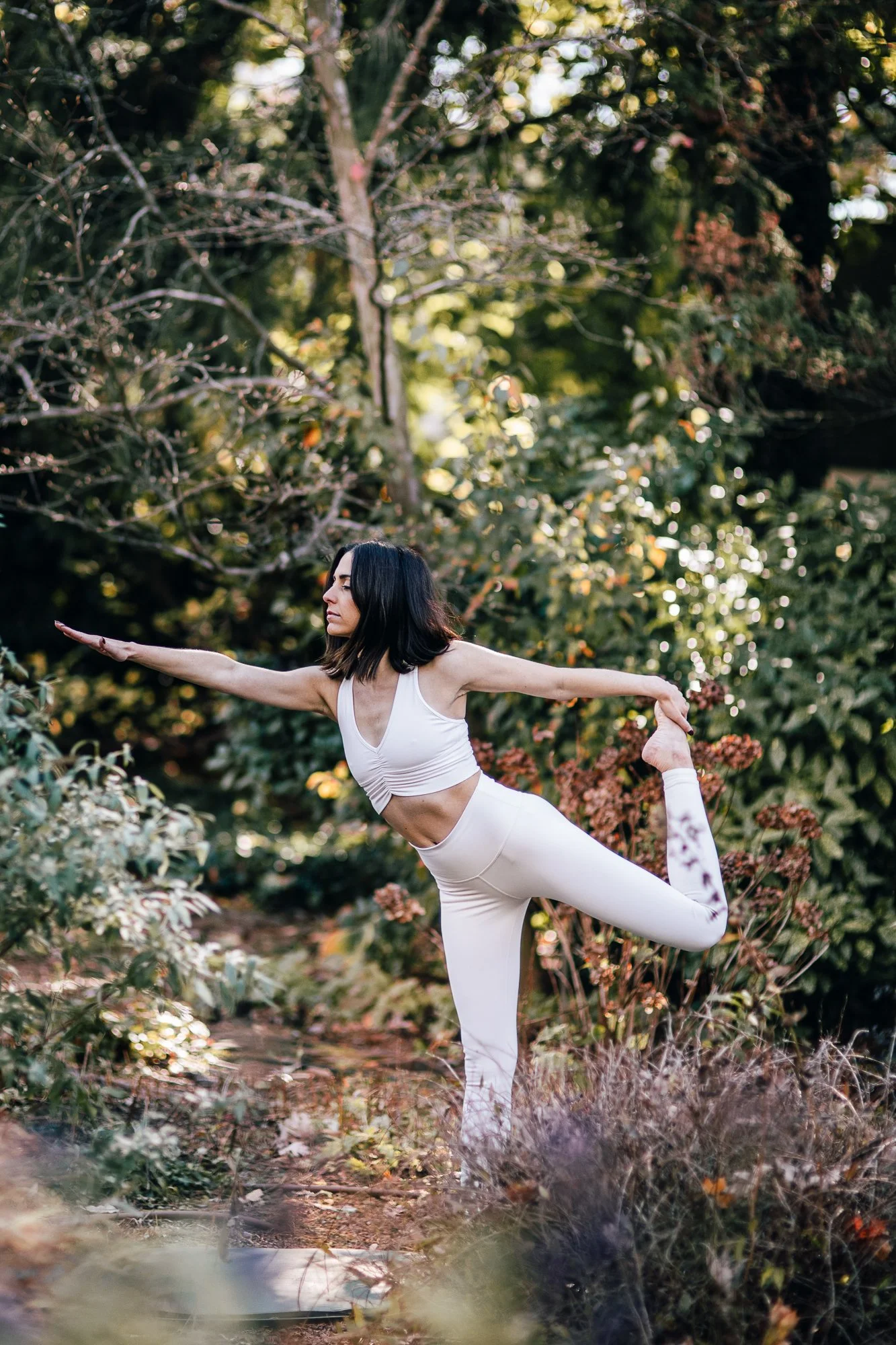 A woman with dark hair practicing yoga outdoors in nature, standing on one leg in a tree pose with arms extended, surrounded by greenery and trees.
