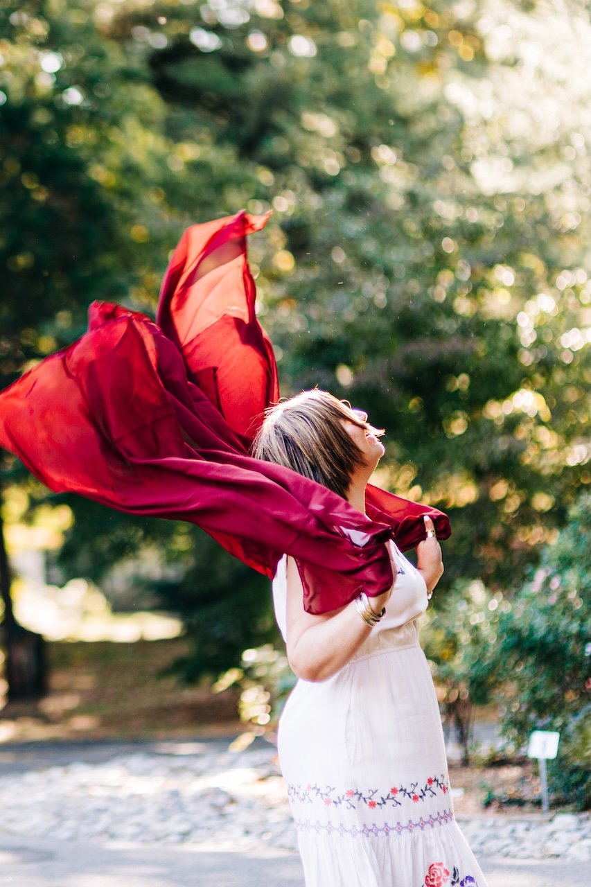 A woman in a white dress holding a flowing red fabric outdoors with trees in the background.