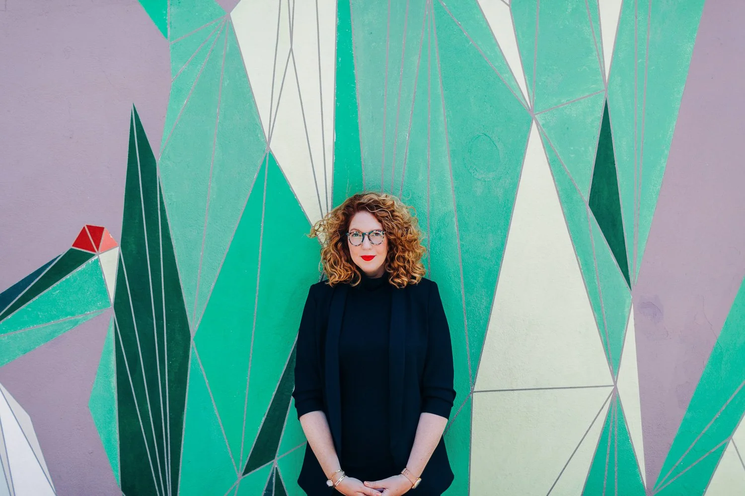 A woman with curly red hair, glasses, and red lipstick standing in front of a colorful geometric mural on a wall.
