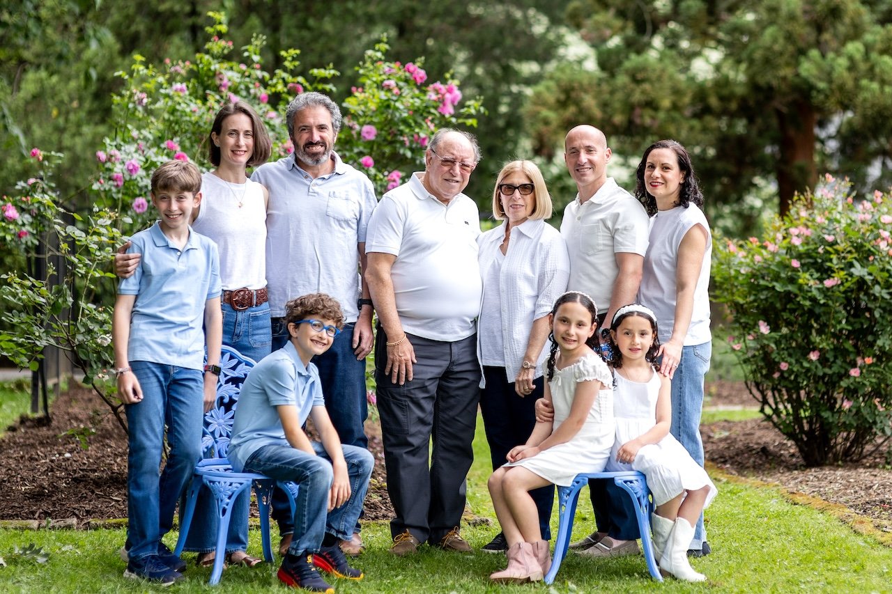 Family of ten people, including children and grandparents, gathered outdoors in a garden with pink flowering bushes behind them. They are smiling and posing for a group photo.
