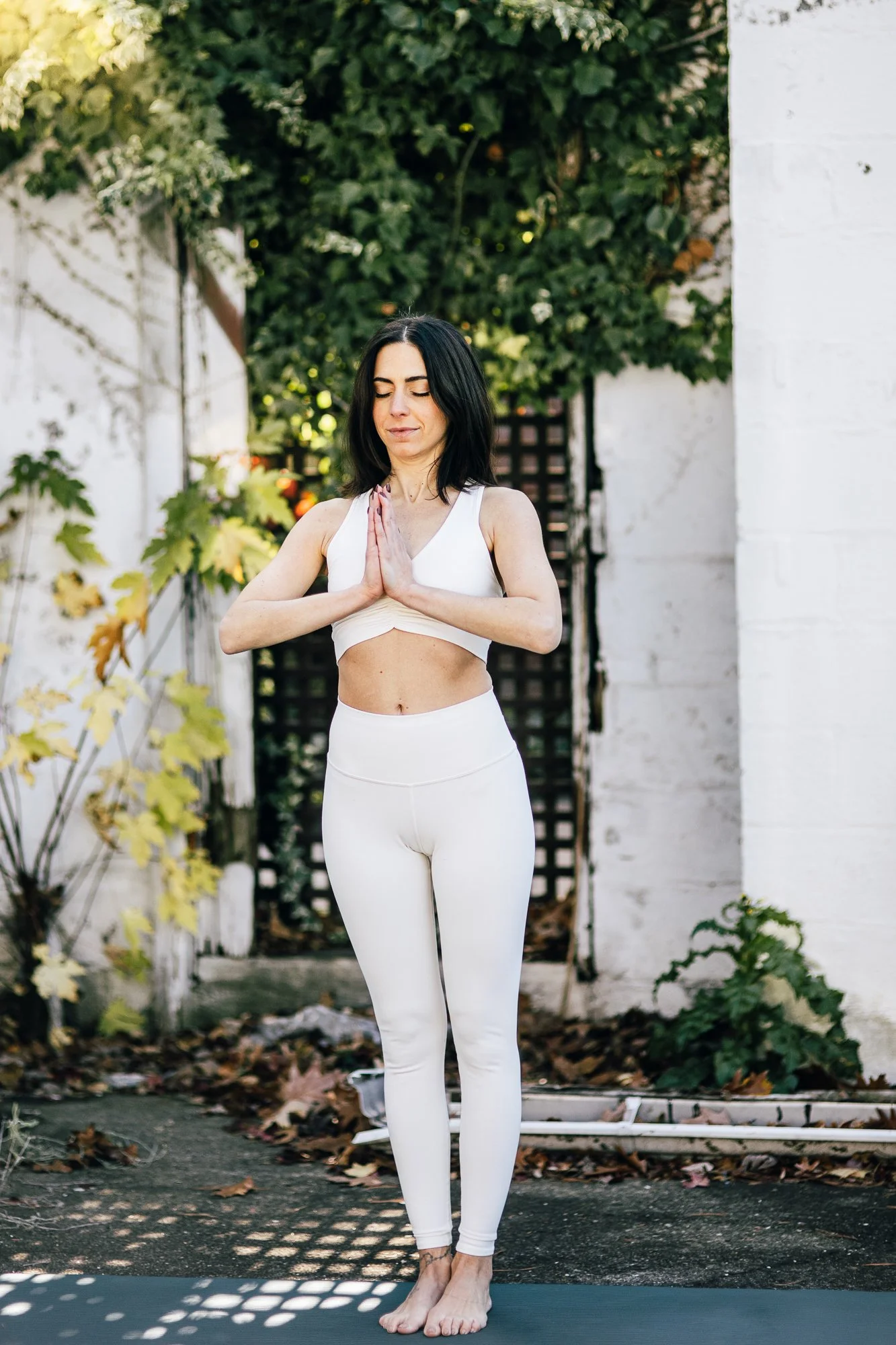 A woman practicing yoga outdoors, standing on a black yoga mat with her hands in prayer position near her chest, eyes closed, dressed in white workout clothes, surrounded by greenery and white walls.