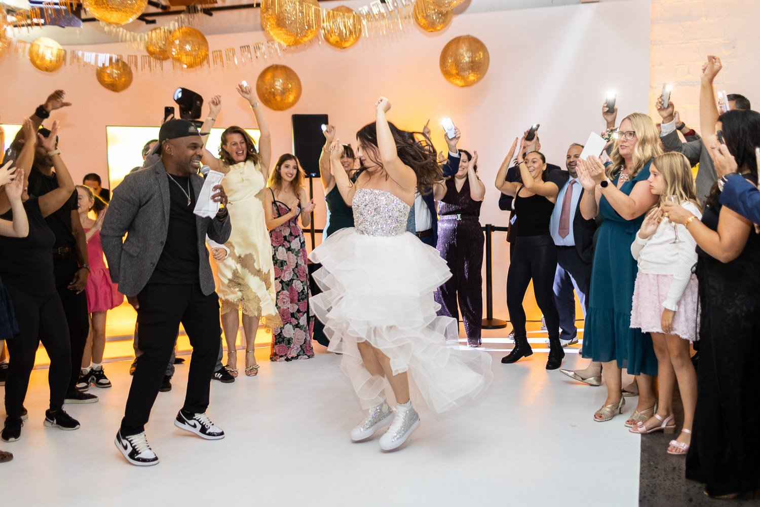 A Bat Mitzvah in a white, ruffled dress and sparkly socks dancing in the center of a party with people surrounding her, some holding up phones to take pictures, in a decorated indoor space with orange balloons hanging from the ceiling.
