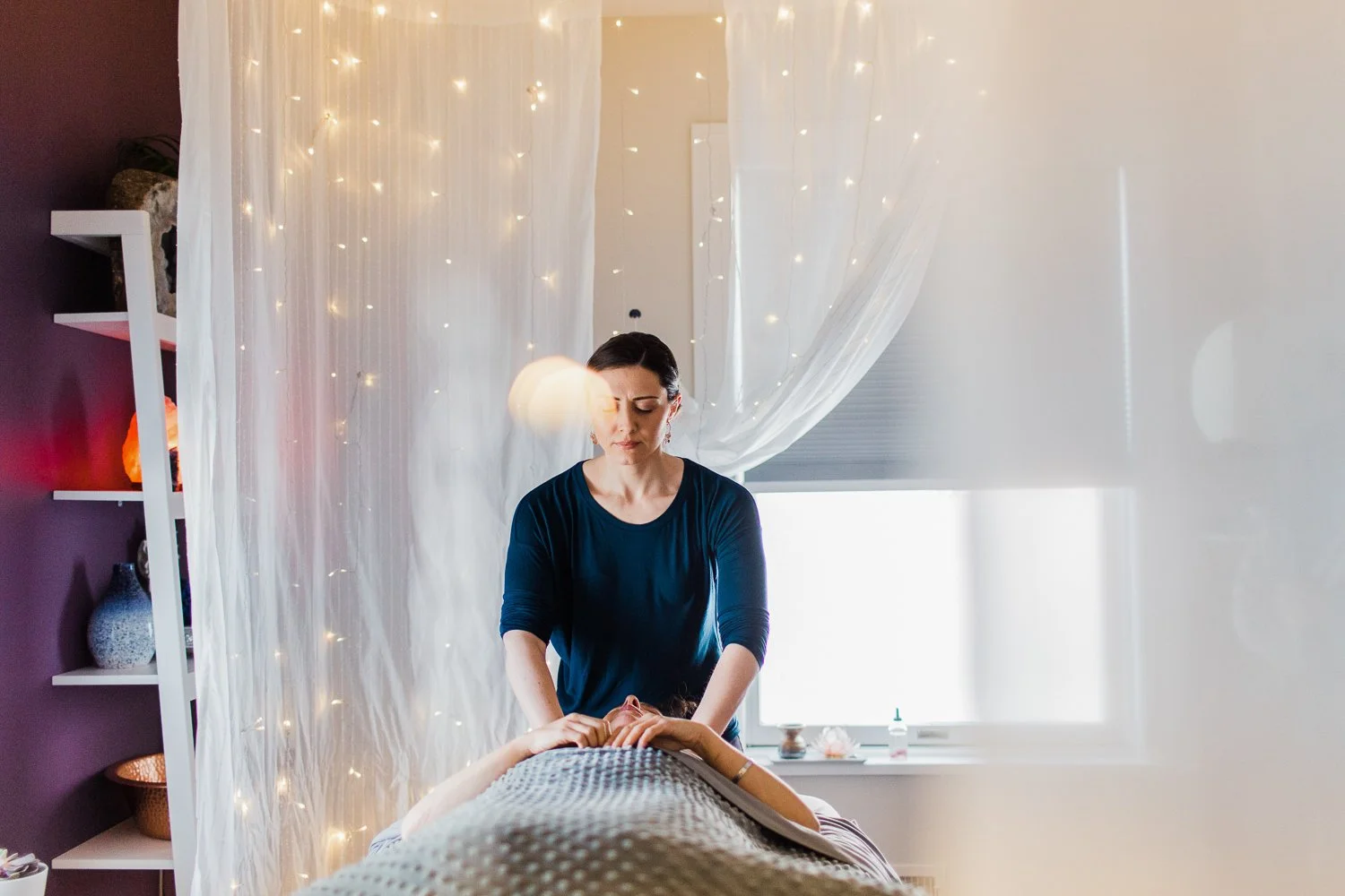 A woman giving a massage to another person lying face up on a massage table in a well-lit, serene room decorated with fairy lights and sheer curtains.