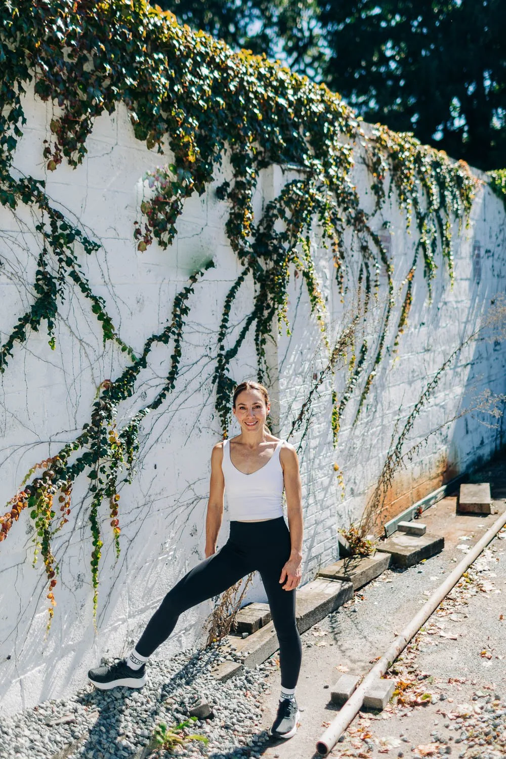 A smiling woman in athletic attire stands confidently with one leg extended against a white wall covered in climbing ivy, outdoors on a sunny day.