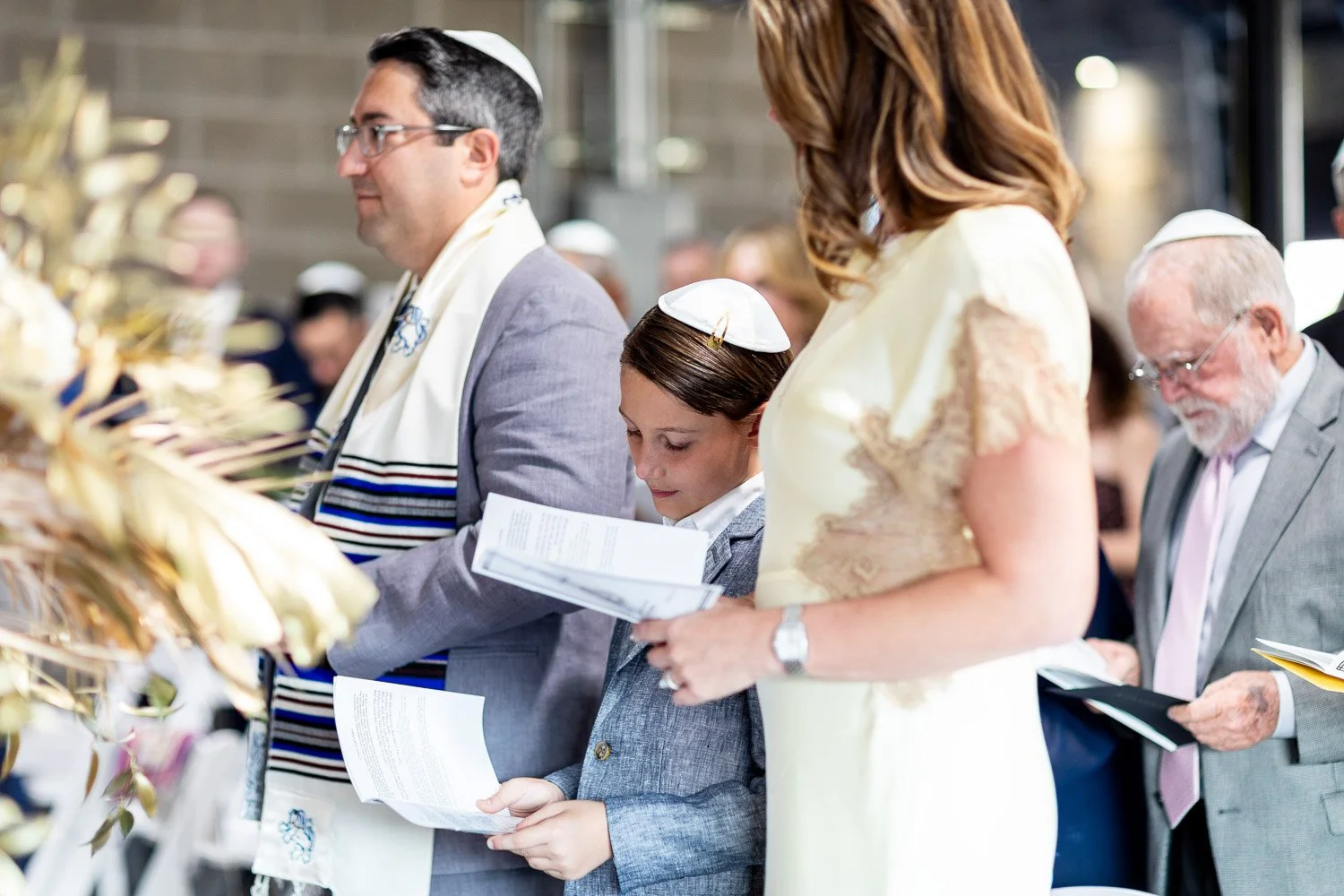People participating in a Bat Mitzvah, some wearing traditional Jewish attire, holding prayer books in a well-lit indoor setting.