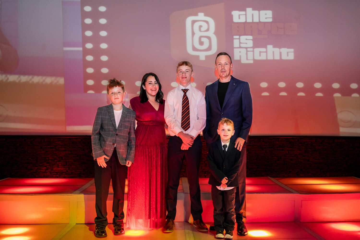 Group of six people, five children and one adult, standing on a stage with a pink background that says "the age is right" and a logo. They are dressed in formal and semi-formal clothing.