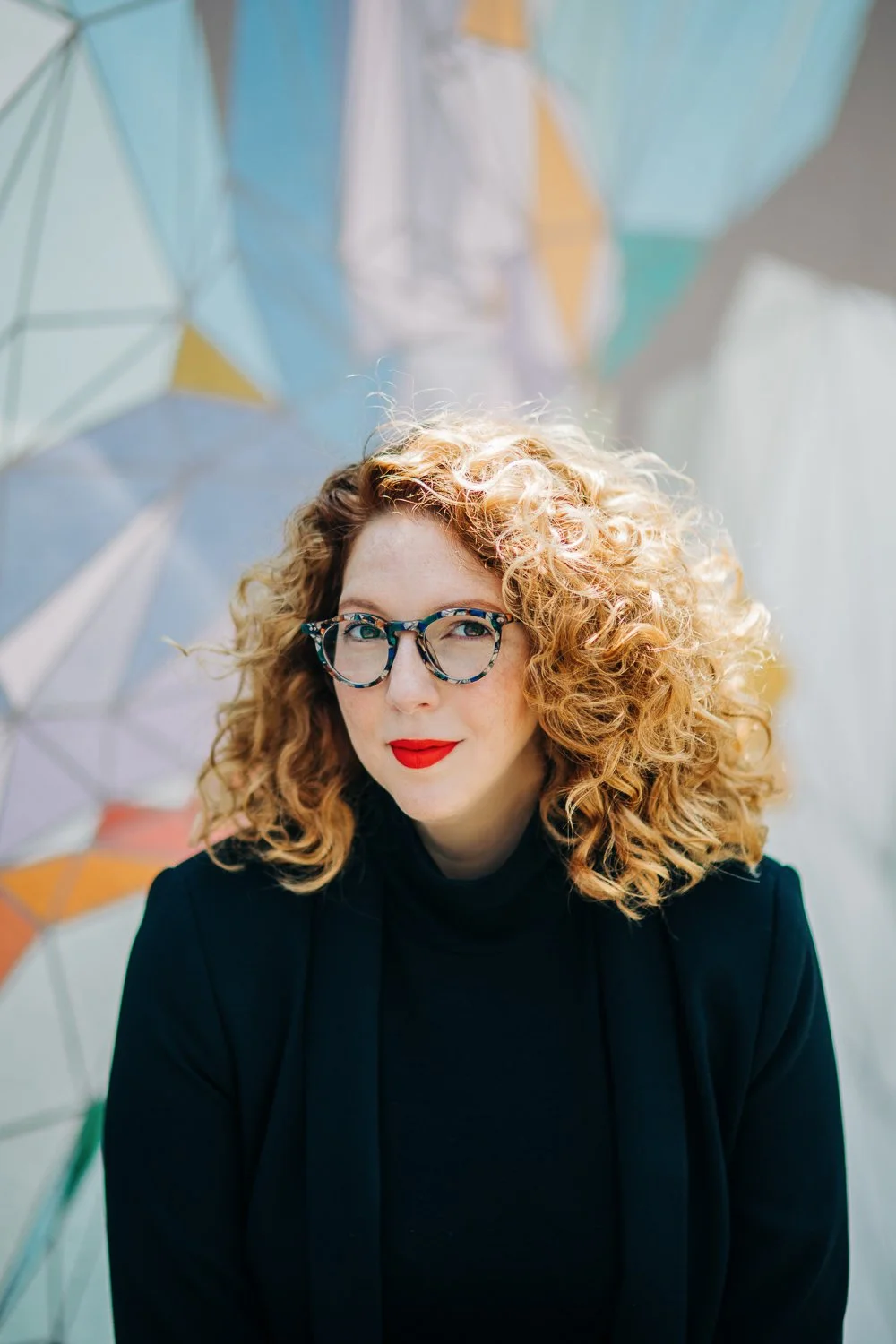 A woman with curly red hair and glasses, wearing a black top and red lipstick, standing in front of colorful umbrellas.