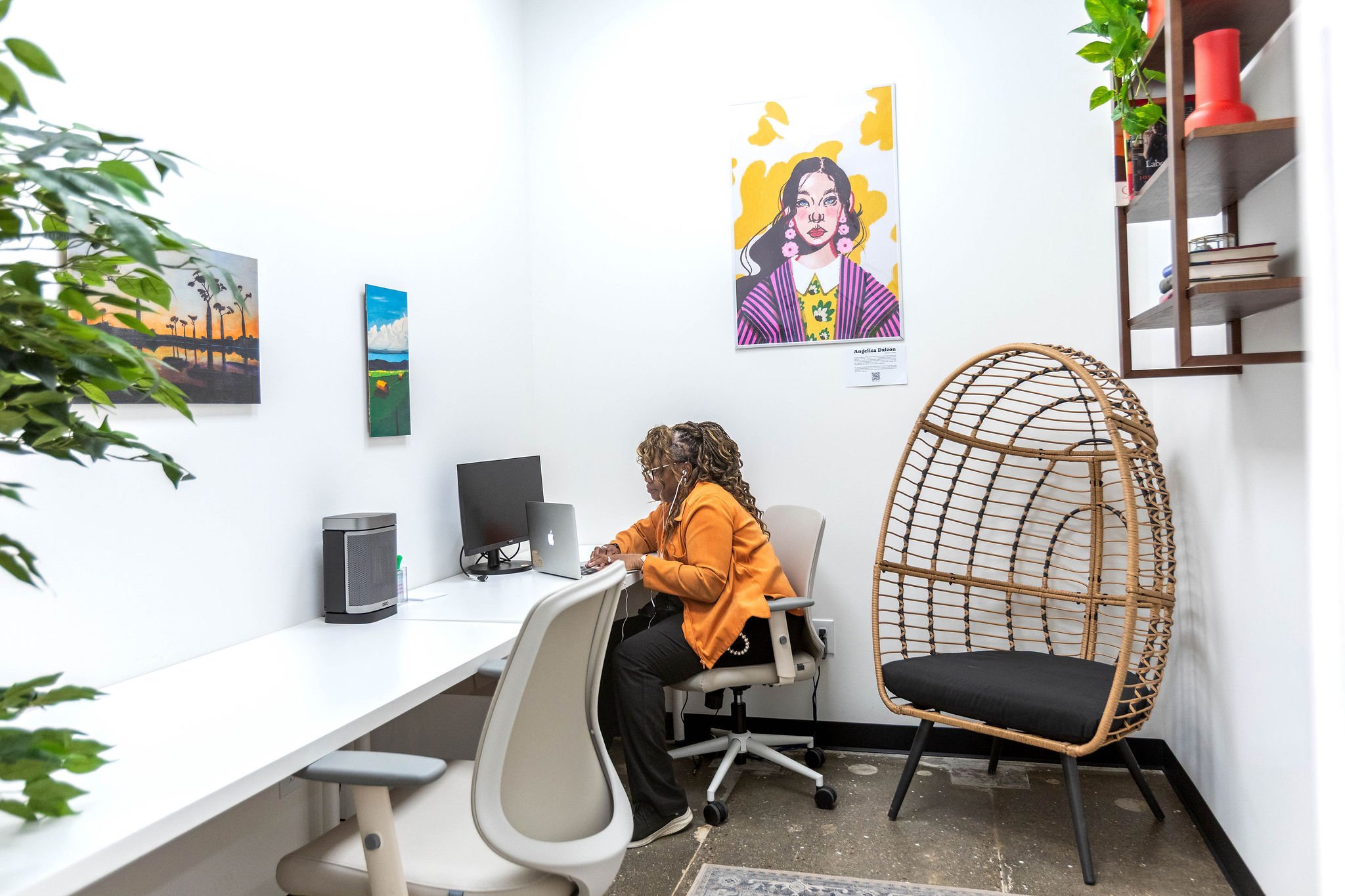 An office with white walls decorated with colorful artwork, a woman with curly hair and glasses working at a desk with a computer, a rattan chair, and potted plants on a shelf.