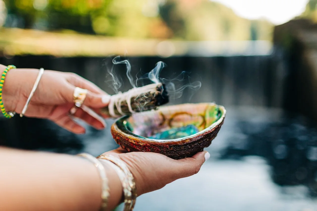 Person holding a steaming, multicolored ceramic bowl with a bundle of burning sage in an outdoor setting.