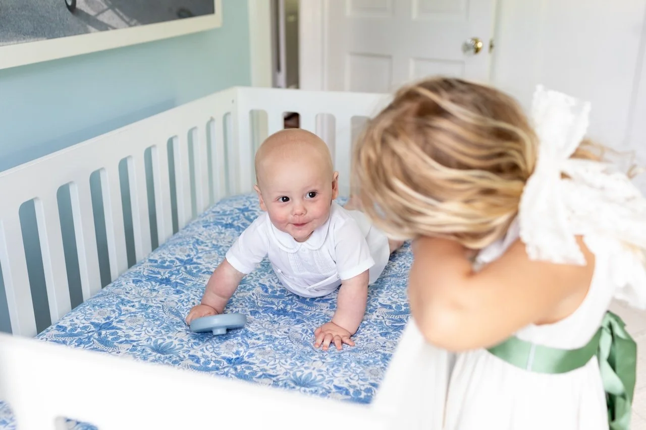 A baby with a white shirt lying on a blue patterned bed, looking at a woman with blonde hair who is leaning over the bed, inside a room with white and light blue walls.