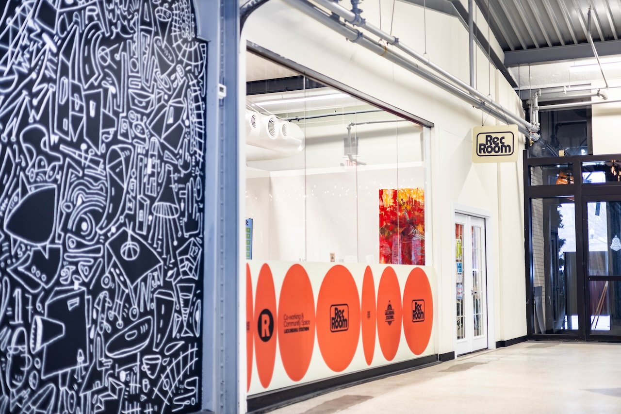 Interior view of a community space with a white wall decorated with orange circles and black text that reads 'Rec Room.' There is a door with glass panels, and a colorful abstract painting is visible inside. A black and white patterned wall with abst