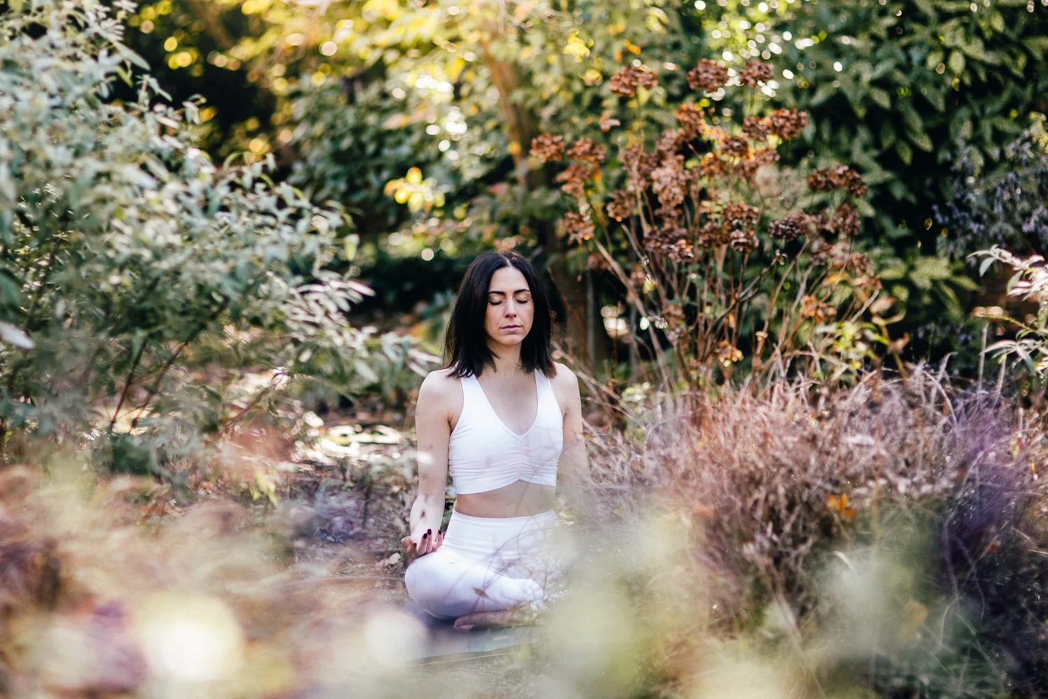 A woman with dark hair in a white sports bra and leggings practicing meditation outdoors surrounded by lush greenery and plants.