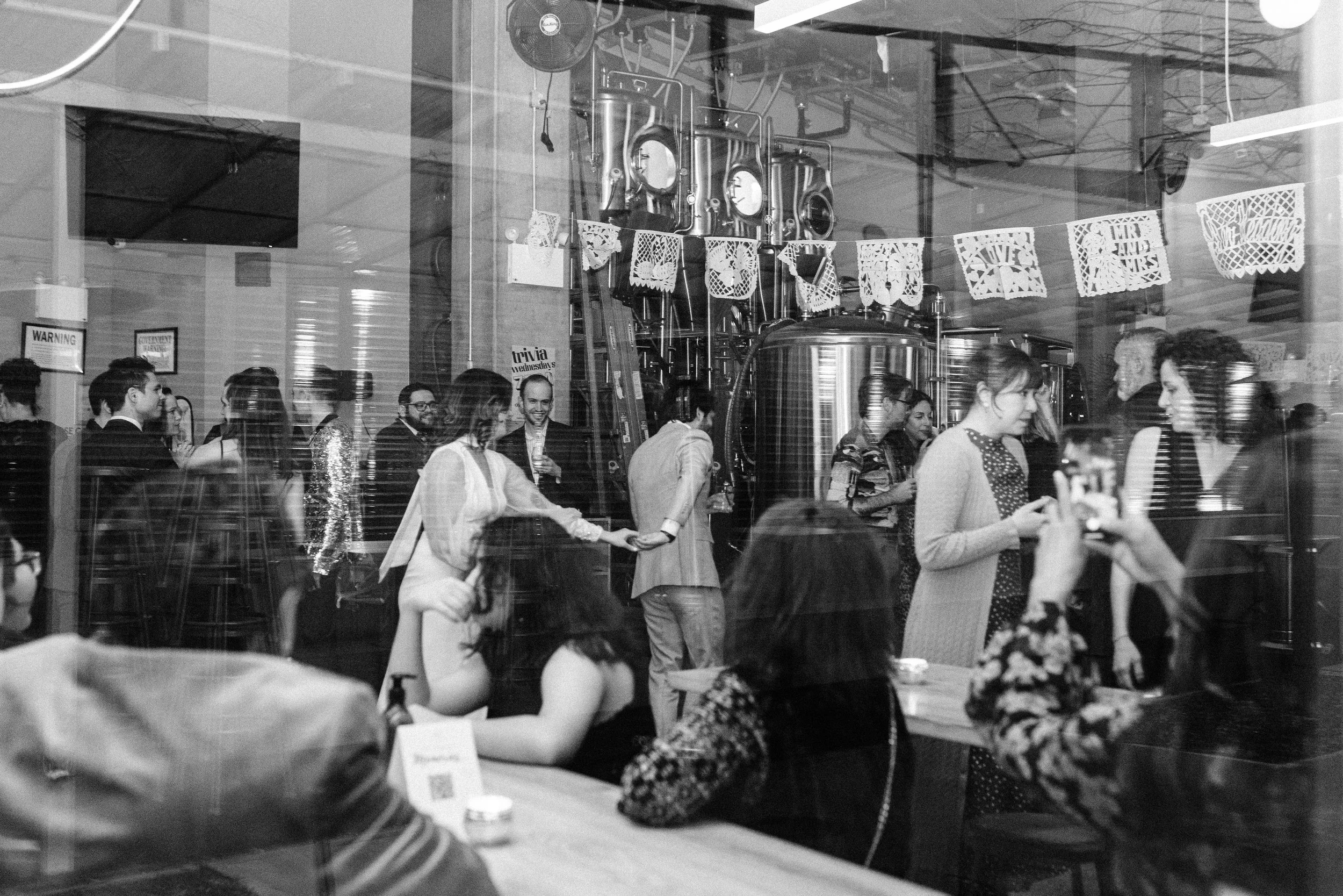 People gathered inside a brewery or cafe, some talking and smiling, with decorated paper banners hanging from the ceiling.