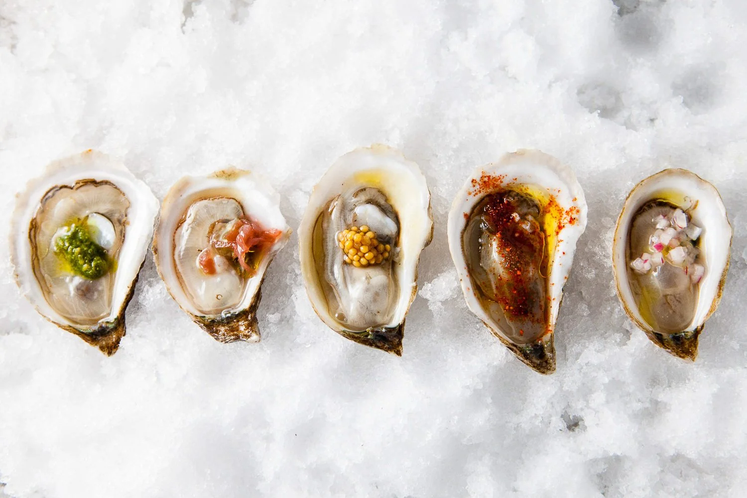 Five raw oysters on a bed of crushed ice, each topped with different sauces and garnishes.