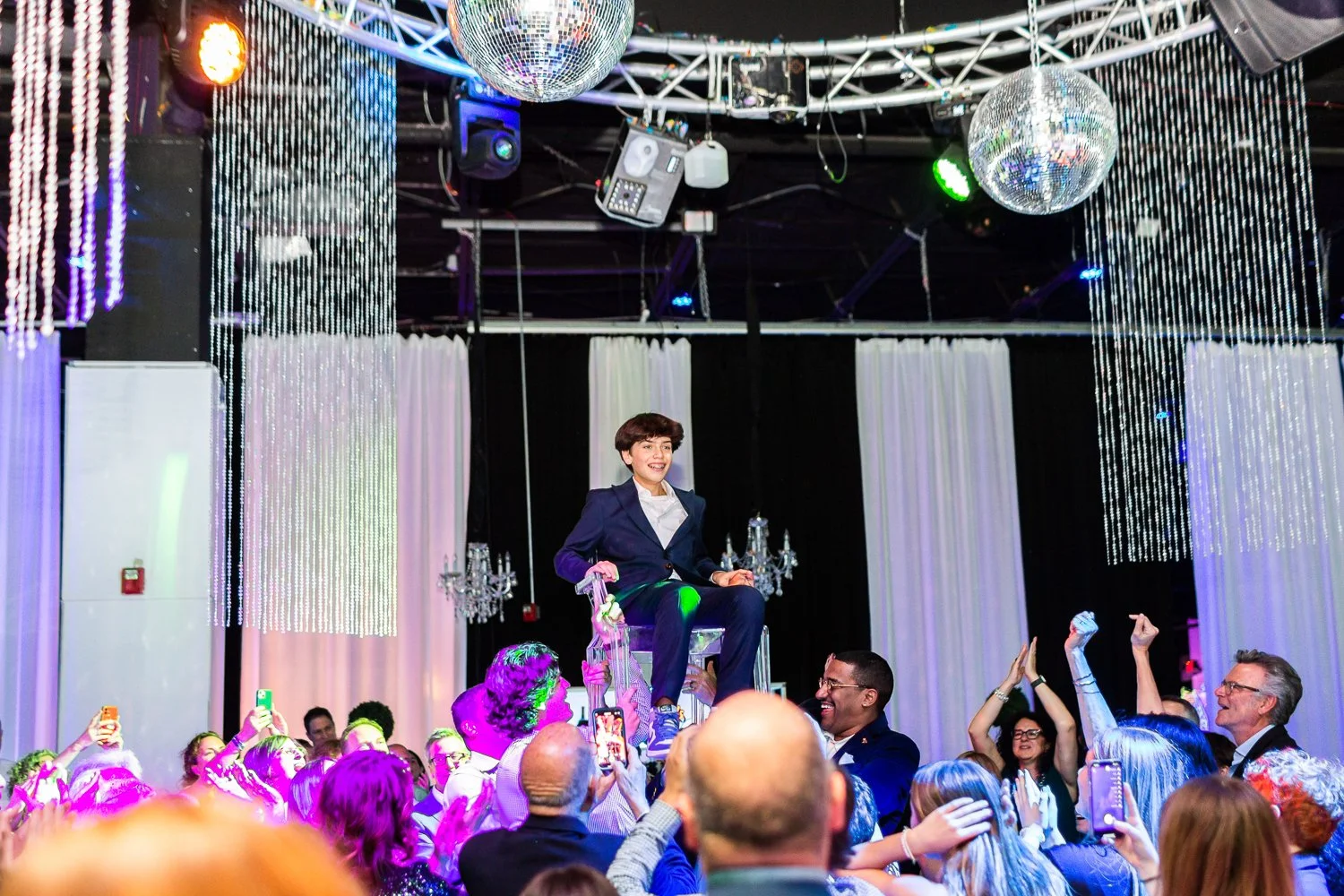 A woman in a navy suit sitting on a throne-like chair lifted above the crowd at a celebration event. The scene is decorated with hanging crystal strands, chandeliers, and disco balls, with colorful lighting and many people taking photos and cheering.