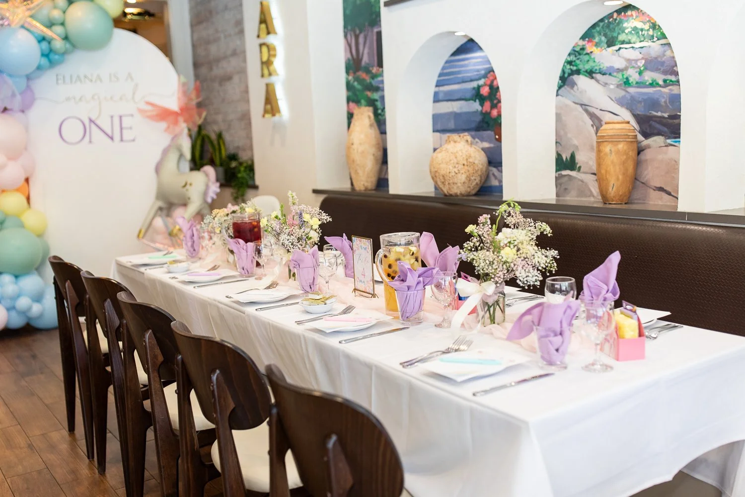 Decorated long dining table with purple napkins, floral centerpieces, and drink pitchers, set for a celebration in a decorated venue.