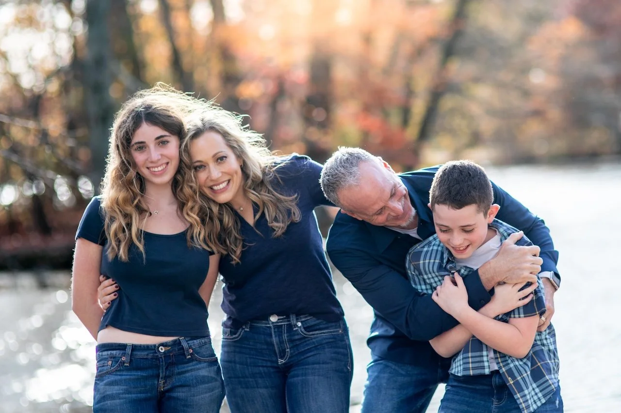 A family of four enjoying time outdoors near a body of water with autumn trees in the background. The father is playfully hugging his young son, while the two women, likely the mother and daughter, smile at the camera. Disability inclusive