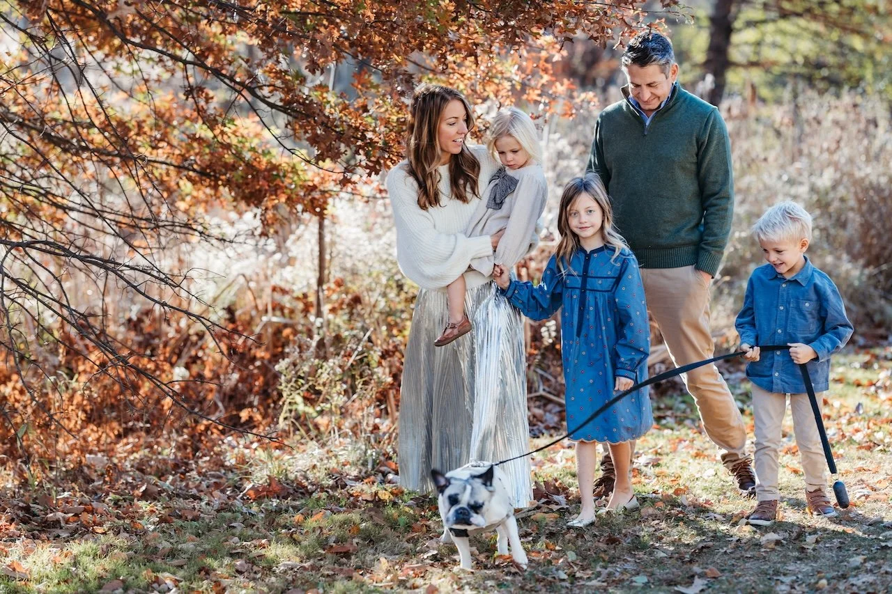 A family of five, including two children, stands outdoors in a park during autumn, with colorful fall foliage around them. They are smiling and interacting, with the children holding a small dog on a leash.
