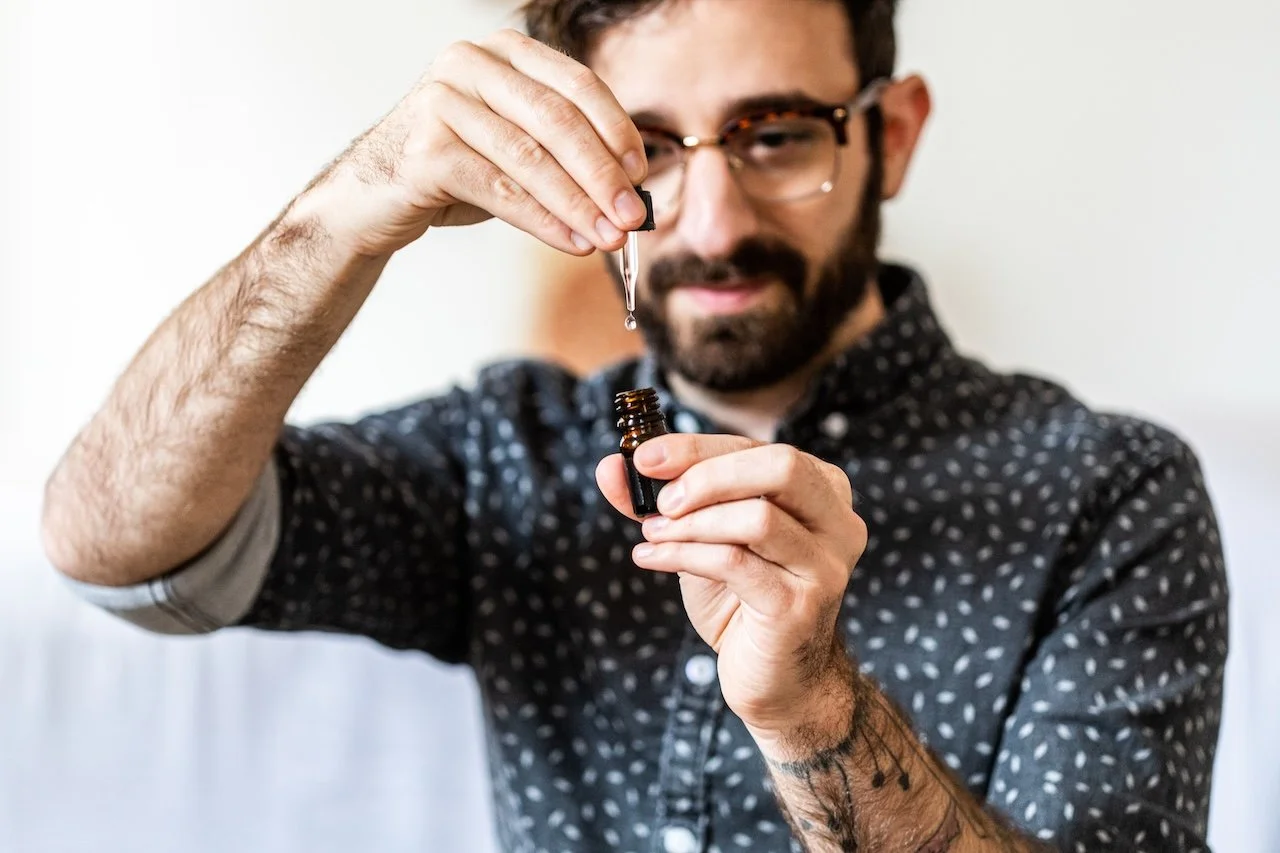 Man with glasses and tattoos using a dropper to transfer liquid into a small brown bottle.