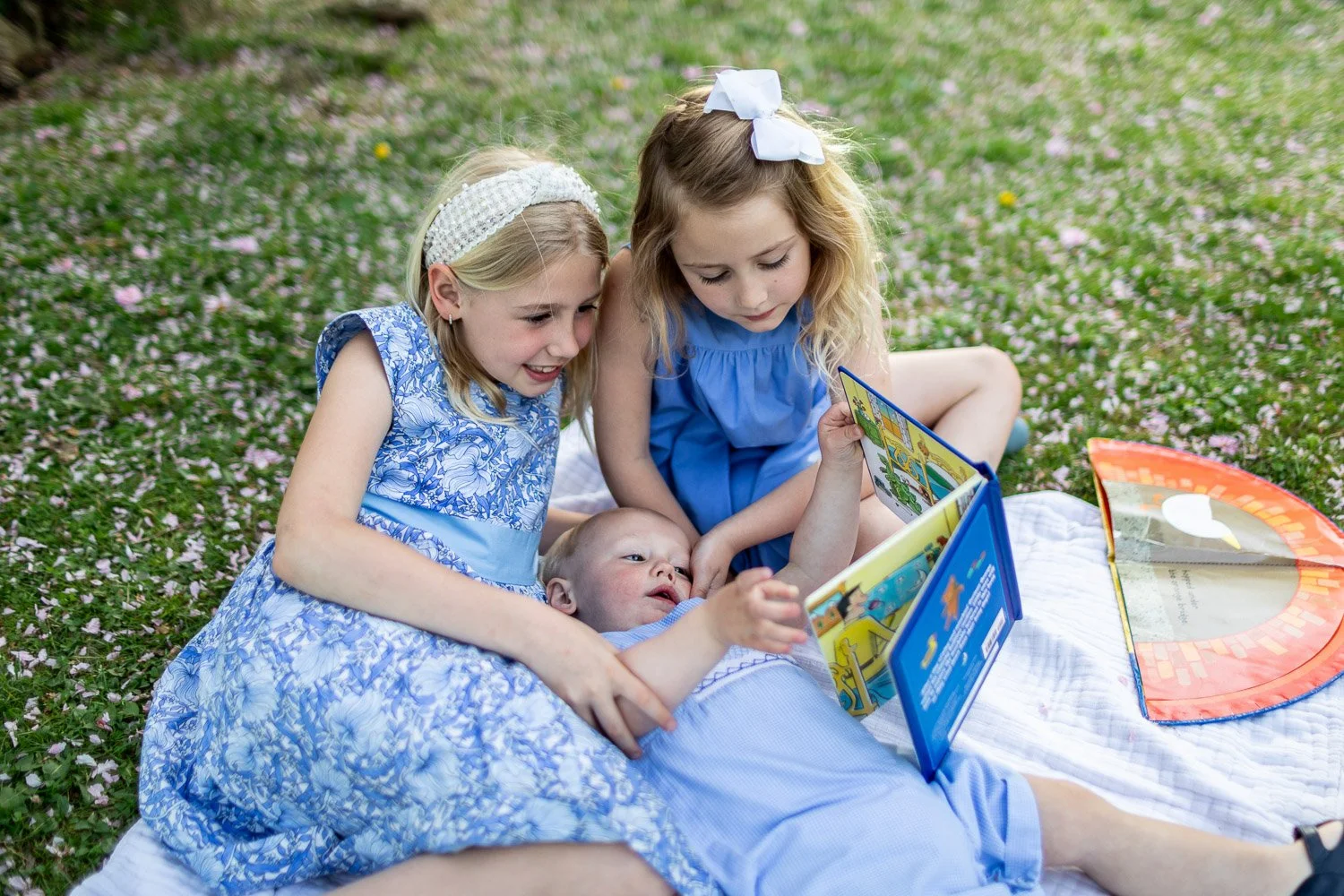 Three young girls and a baby lying on a white blanket on the grass, looking at a colorful book together