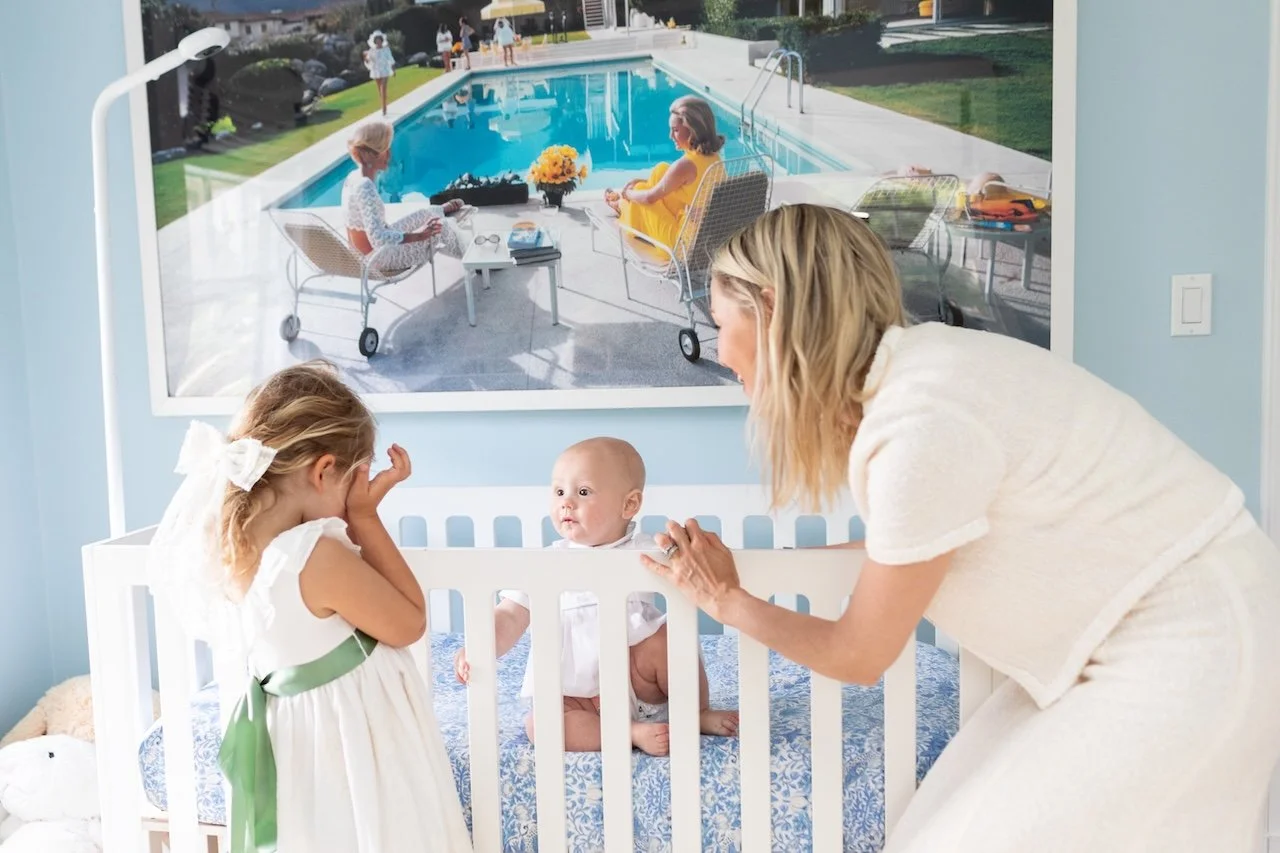 A woman and two young girls with a baby in a crib in a room, with a large window showing an outdoor swimming pool scene with people relaxing by the pool.