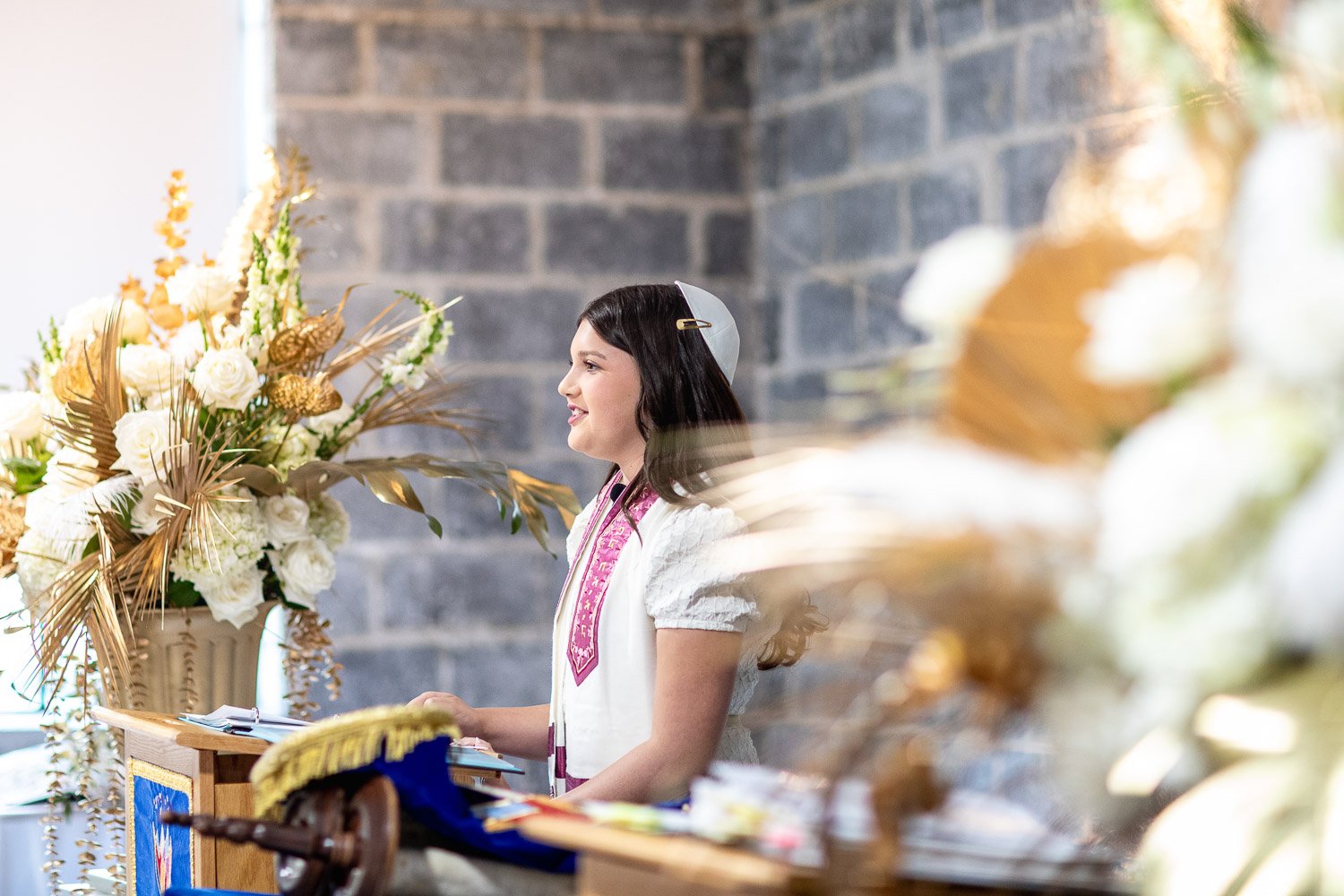 A young woman wearing a white dress and pink ribbon standing at a wooden altar with a large floral arrangement of white and gold flowers and foliage, indoors with a brick wall background.