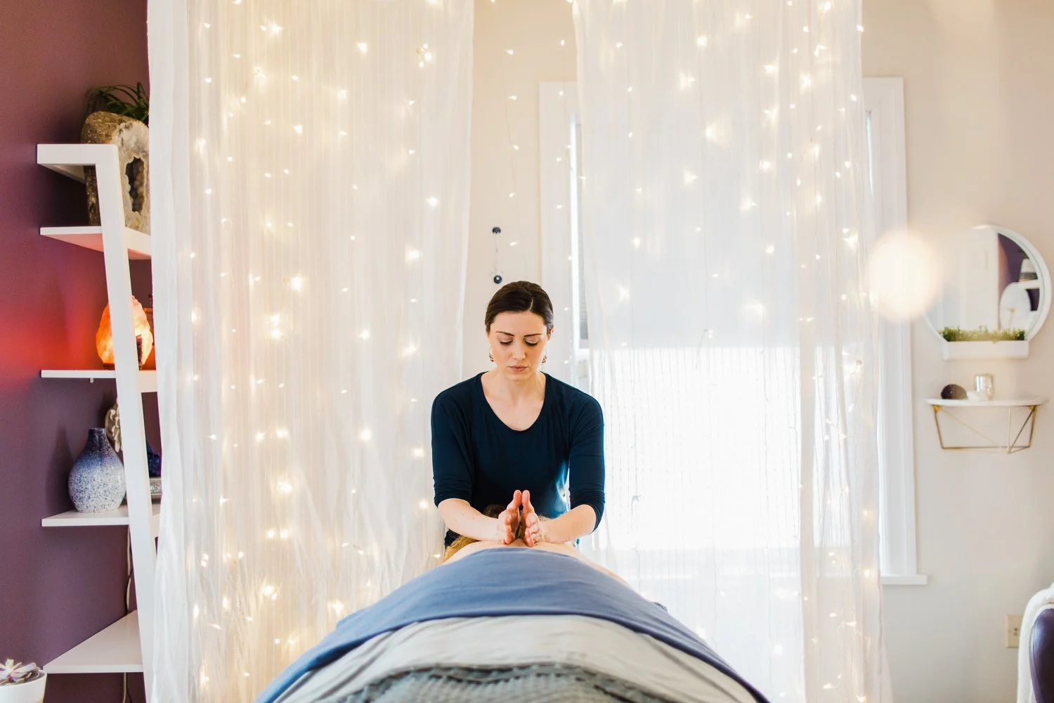 A woman gives a massage to a person who is lying face down on a massage table in a room with white curtains and string lights, with decorative shelves on the wall. Branding photo for masseuse and birth doula.