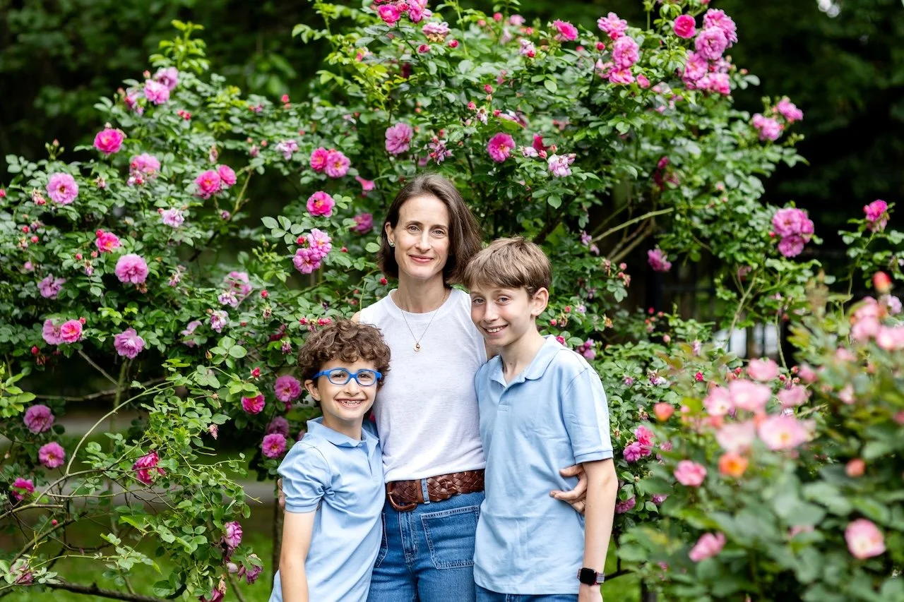 A woman and two boys stand outdoors in front of a large bush with pink roses. All three are smiling and looking at the camera.