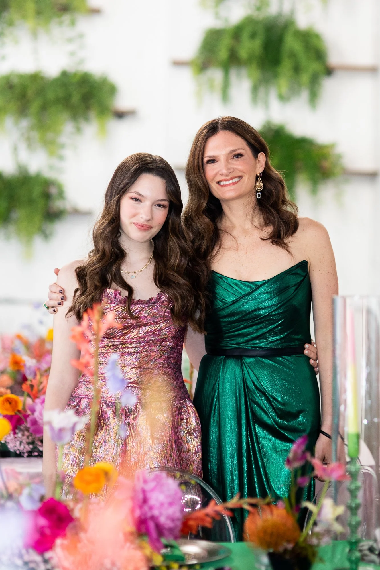 A woman and a young girl standing together at a table filled with colorful flowers, smiling at the camera. The woman is wearing a green strapless dress and earrings, and the girl is wearing a pink and metallic dress with necklaces. In the background,