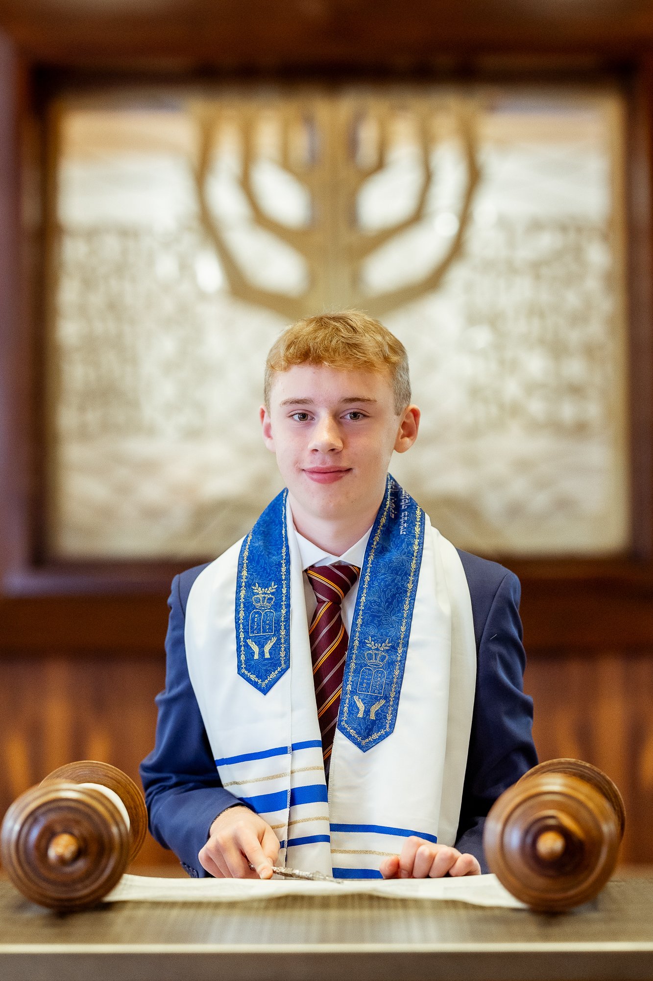 A young man dressed in formal attire, wearing a blue and gold ceremonial stole and a striped tie, is standing at a wooden podium with a Torah scroll in front of him, inside a synagogue. A large shadow of a menorah is cast on the wall behind him.