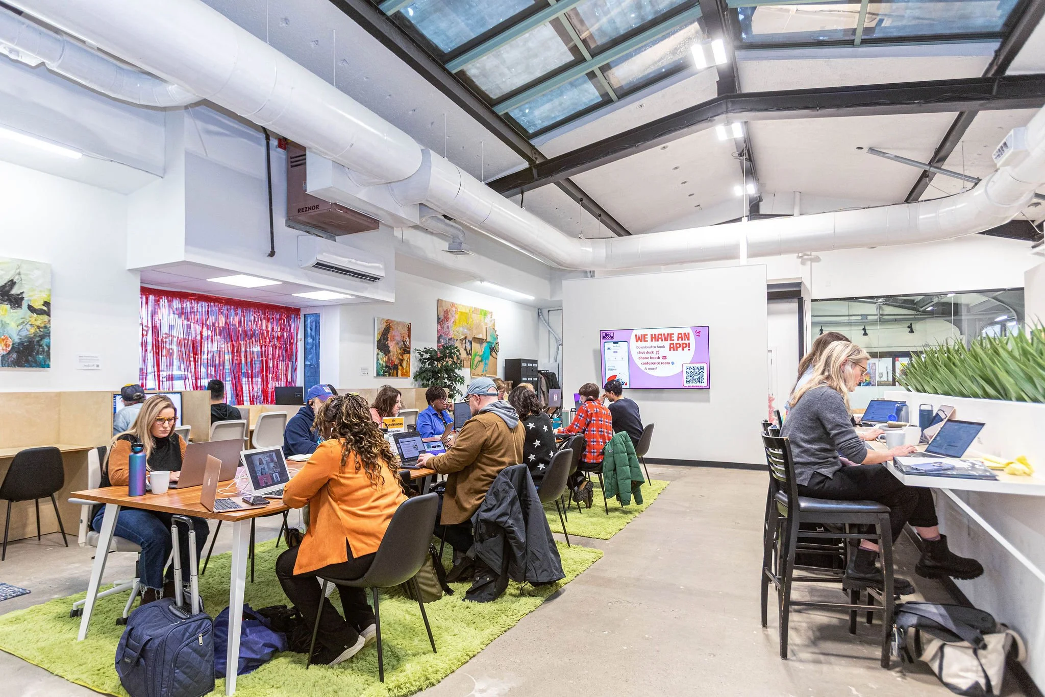 A coworking space with people working on laptops at tables. There are colorful paintings on the wall, a large window with a metal frame on the ceiling, and a digital sign with a QR code and a message about having an app.