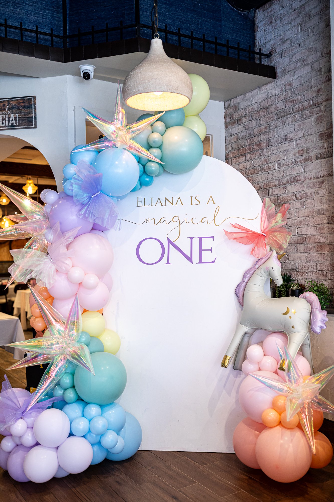Decorative rainbow balloon arch for a first birthday celebration with pastel-colored balloons, iridescent star balloons, a unicorn balloon sculpture, and a white circular backdrop with text saying "Eliana is a magical one."