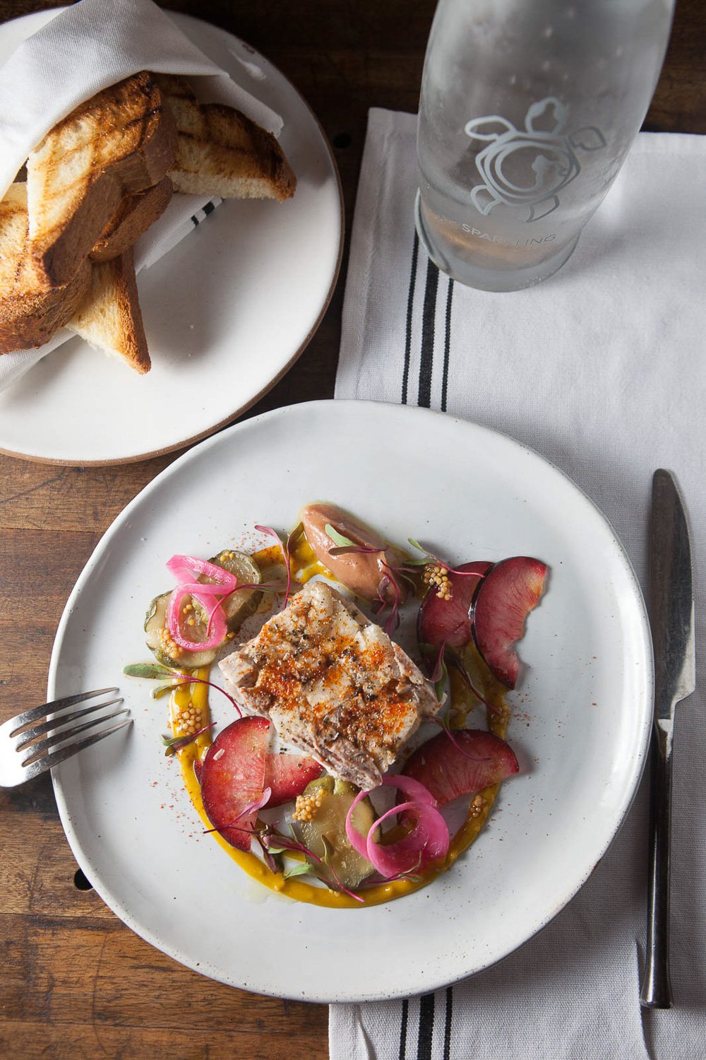 A plate of gourmet food with a serving of fish topped with spices, surrounded by pickled vegetables, and garnished with colorful microgreens and sauces, on a white plate. A basket of toasted bread slices is in the background. A glass of water with a 