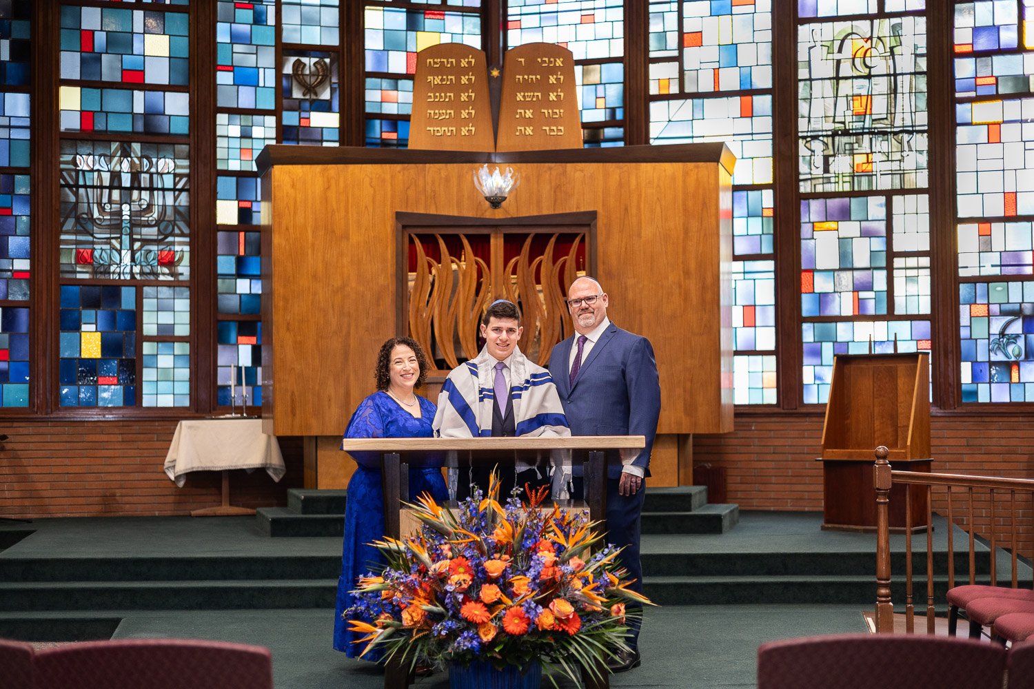 A Jewish bat mitzvah ceremony inside a synagogue with stained glass windows. The bat mitzvah girl, dressed in a white and blue tallit, stands between a woman in a blue dress and a man in a suit, with a floral arrangement in front. The background feat