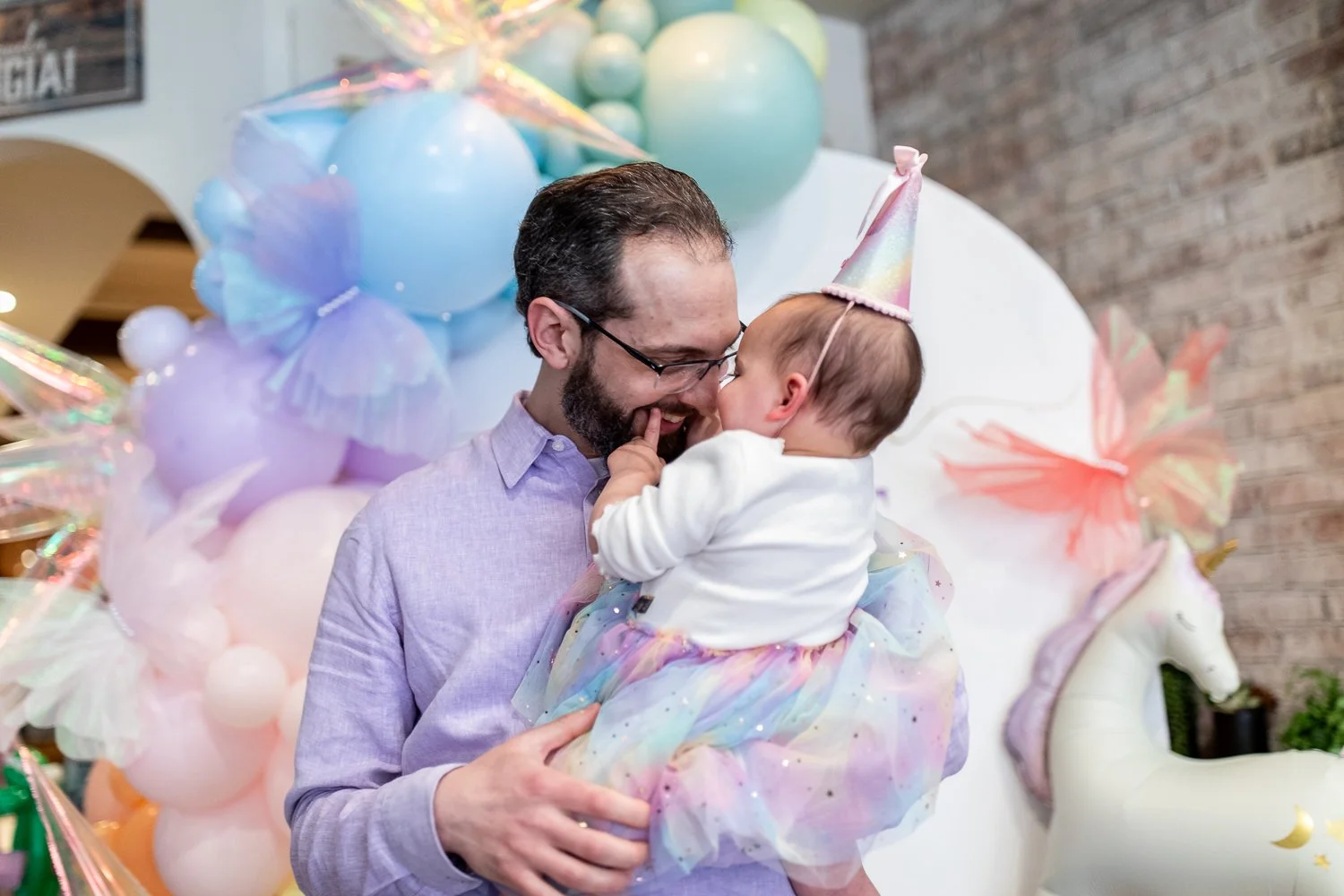 A father and daughter at her 1st birthday celebration in Montclair, NJ share a joyful moment. The girl wears a rainbow-colored tutu and a unicorn-themed party hat, while her dad holds her.
