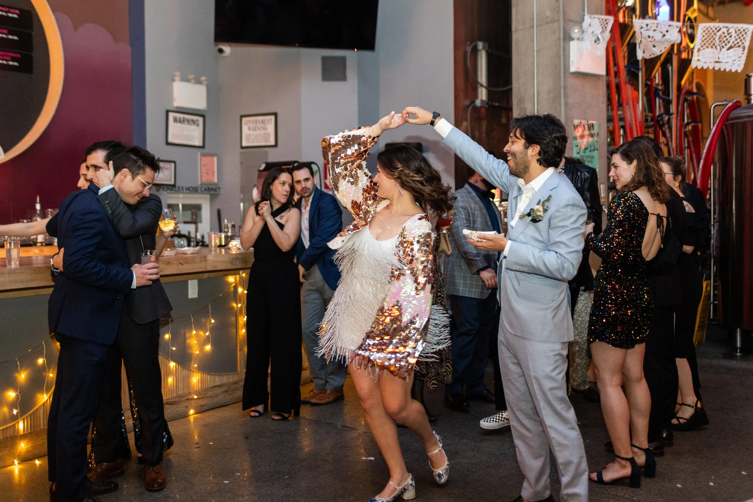 People dancing and socializing at a party or celebration, with a woman and man holding hands and spinning, dressed in formal attire, inside a decorated venue with dim lighting.  This party is taking place in Brooklyn, NY.