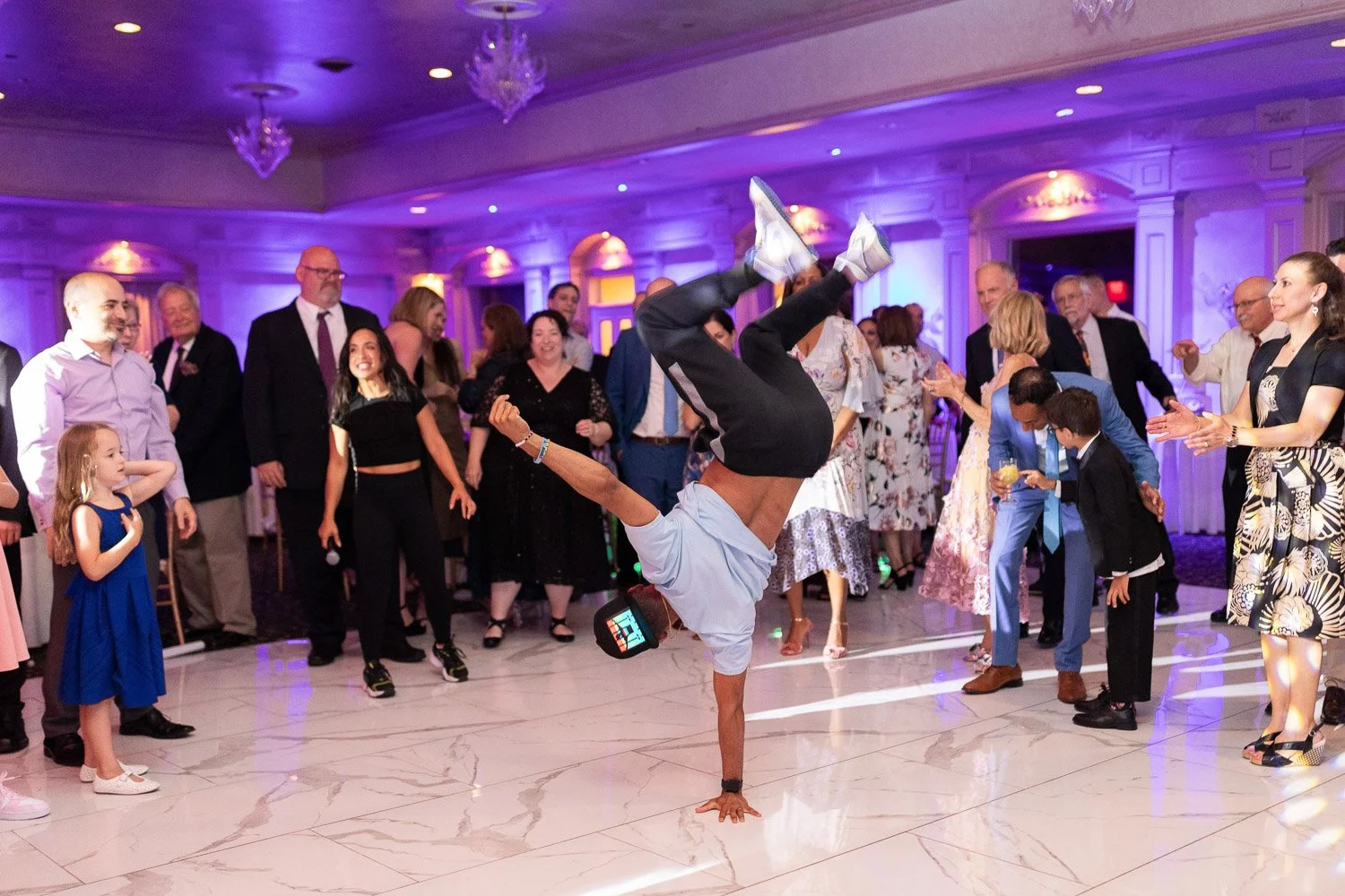 A man performing a handstand in the middle of a dance floor at a party, surrounded by smiling guests clapping and watching. The room has purple lighting and ornate chandeliers.