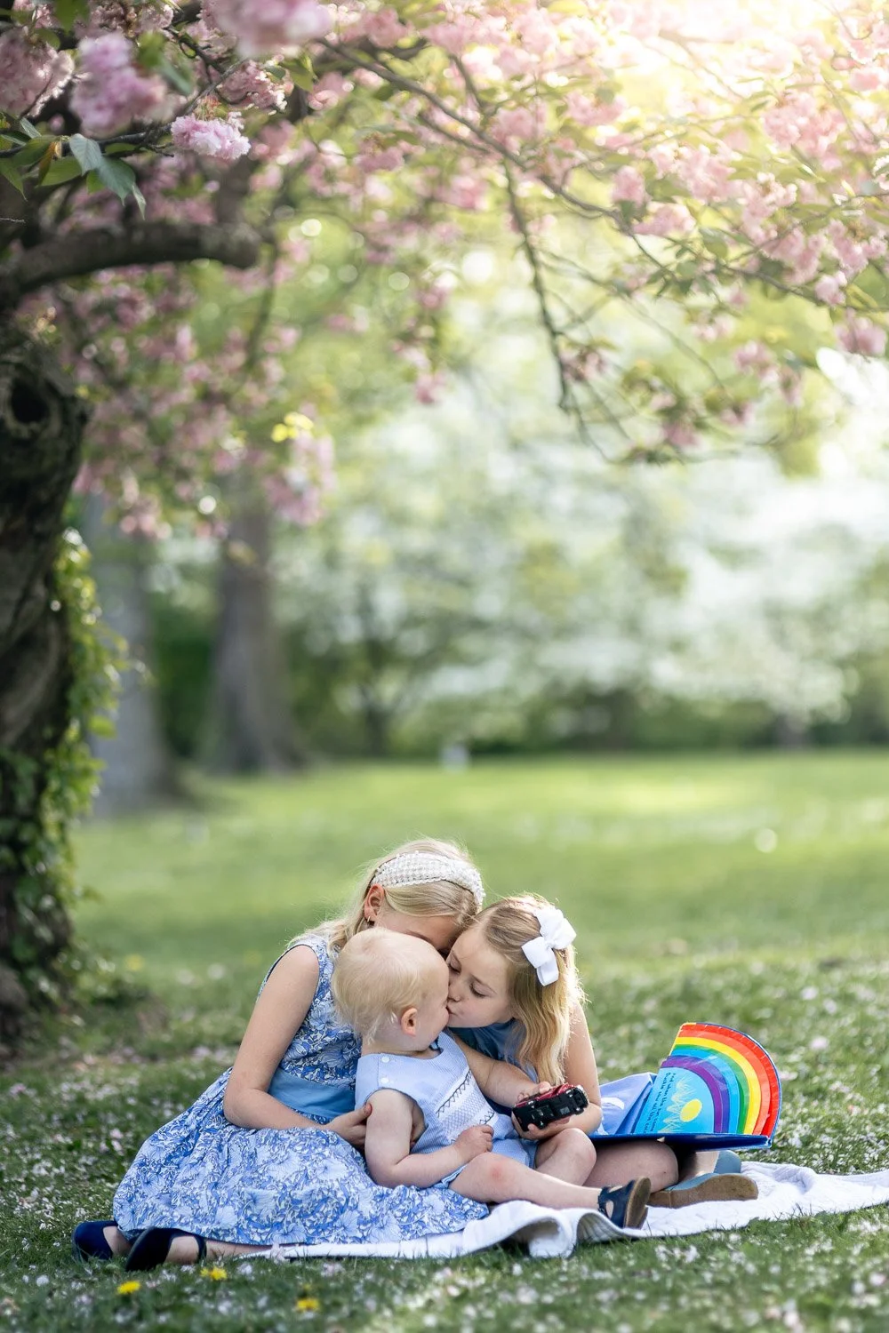 Three young girls sitting on a white blanket on the grass beneath a blooming pink cherry blossom tree, sharing a kiss and enjoying a picnic in a park.
