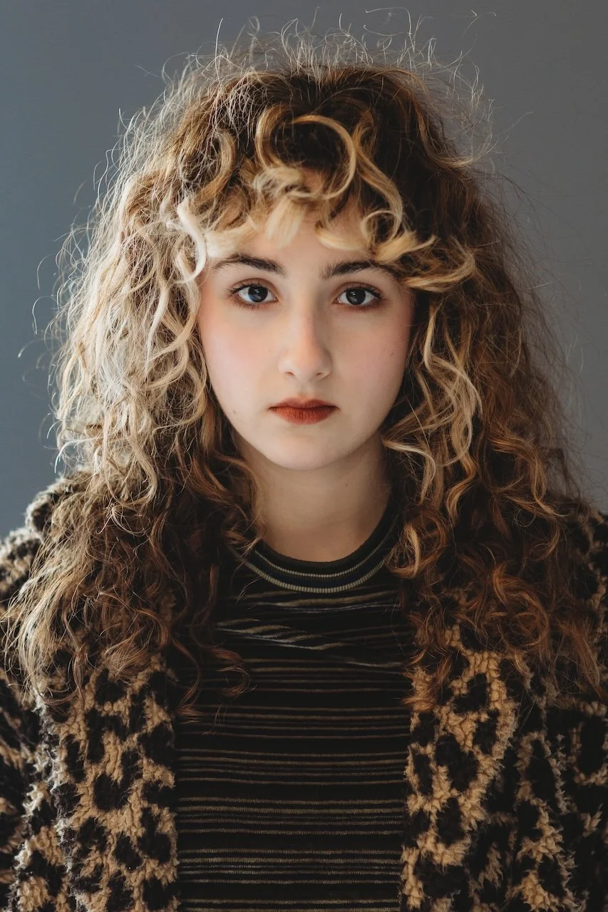 Portrait of a young woman with curly blonde hair, wearing a leopard print coat and a black striped shirt, against a gray background.