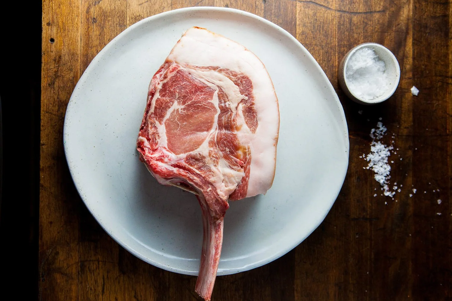 A raw T-bone steak on a white plate, with a small container of coarse sea salt and a small pile of salt on a wooden surface.
