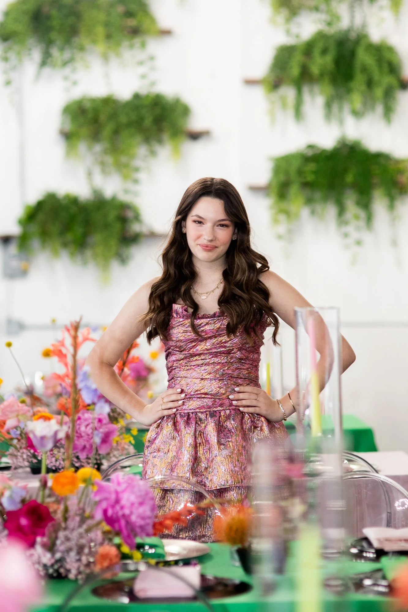 A young woman with long wavy dark brown hair wearing a sleeveless pink and purple patterned dress, standing with hands on hips in front of a colorful flower arrangement at her Bat Mitzvah in NJ.