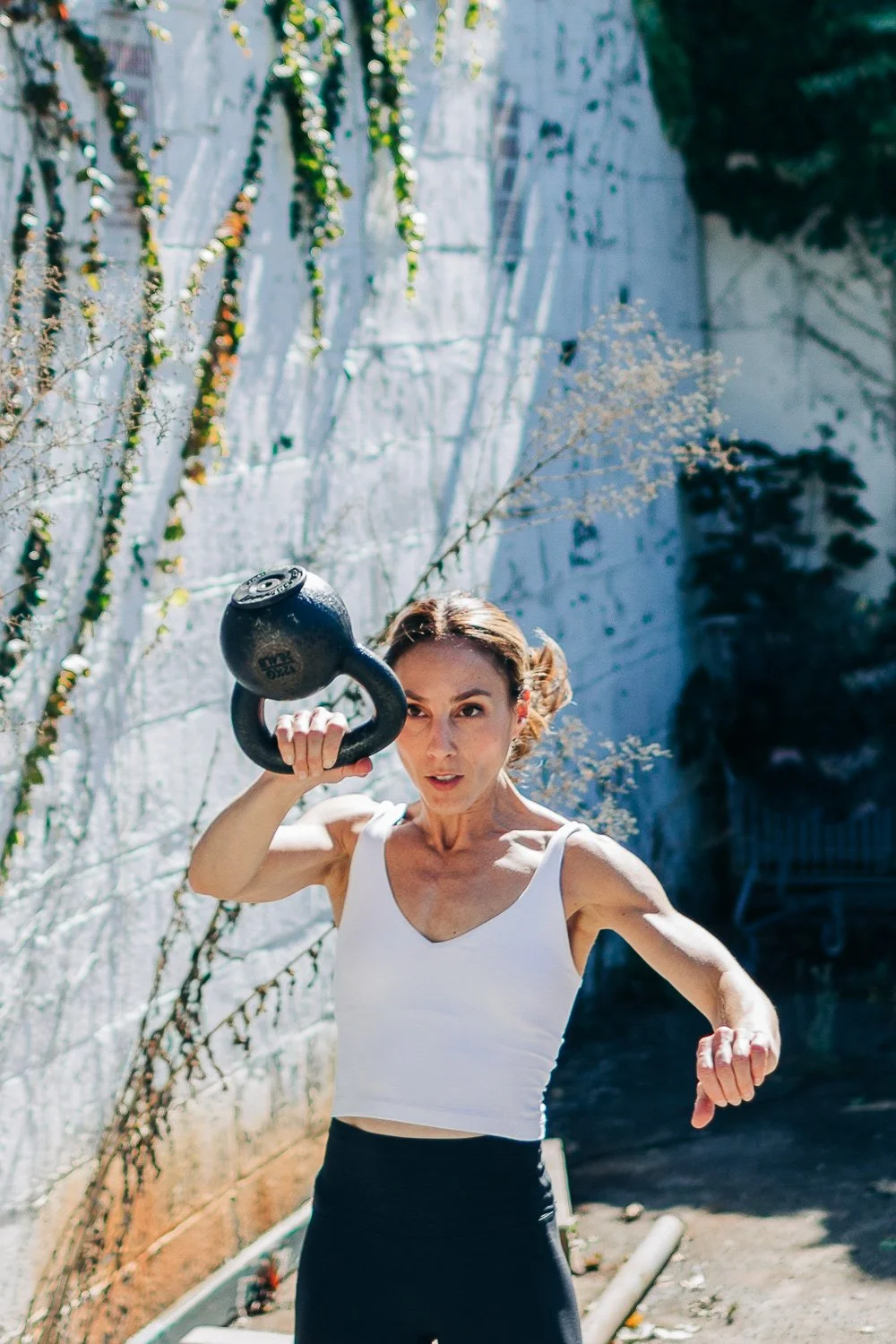 A woman in workout attire holding a kettlebell in an outdoor setting with a white brick wall and greenery.