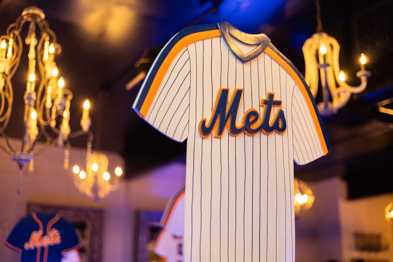A miniature replica of a New York Mets baseball jersey hanging indoors, with chandeliers and seating area visible in the background.