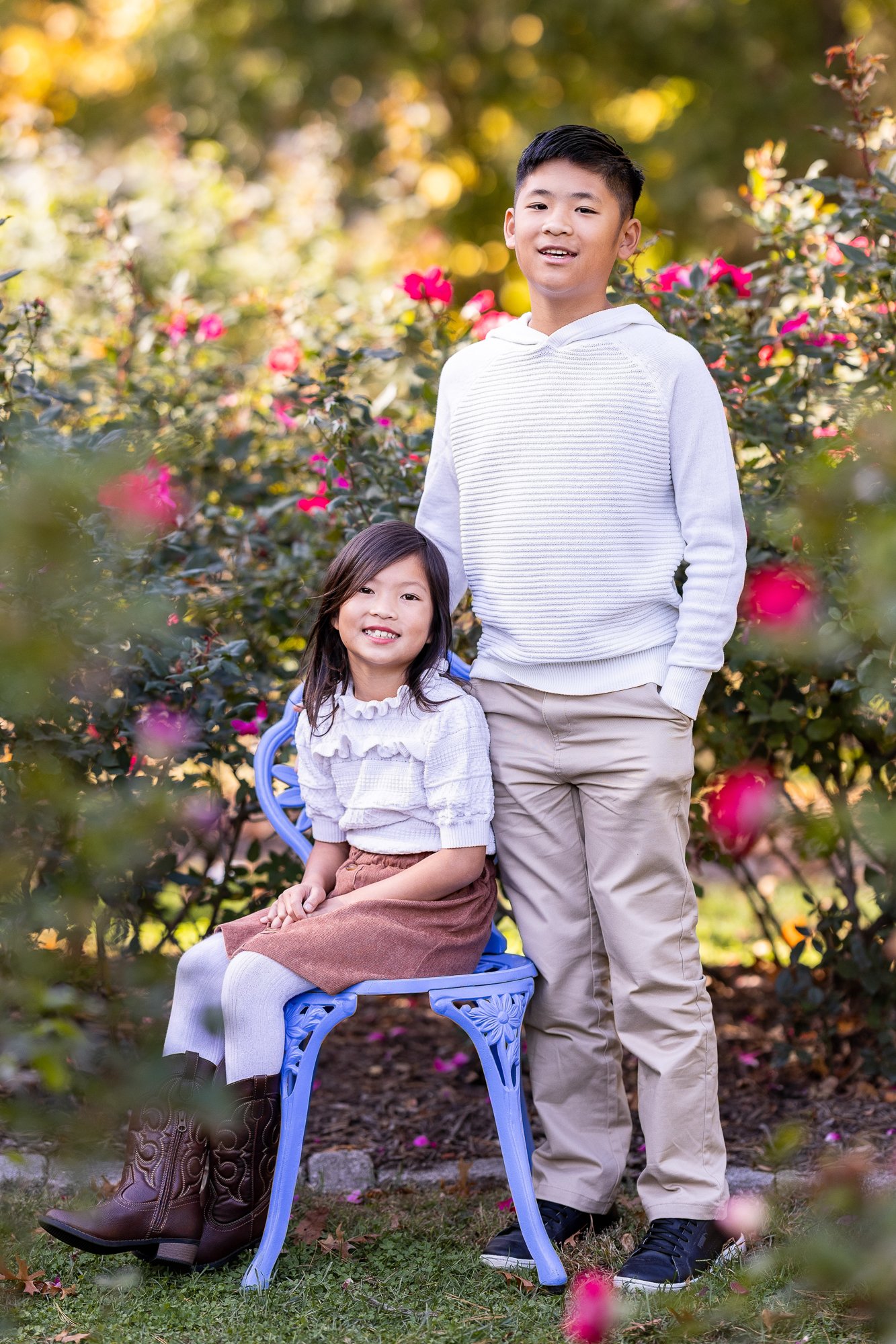 A boy standing beside a girl sitting on a blue park bench, surrounded by pink and yellow flowers and green foliage.