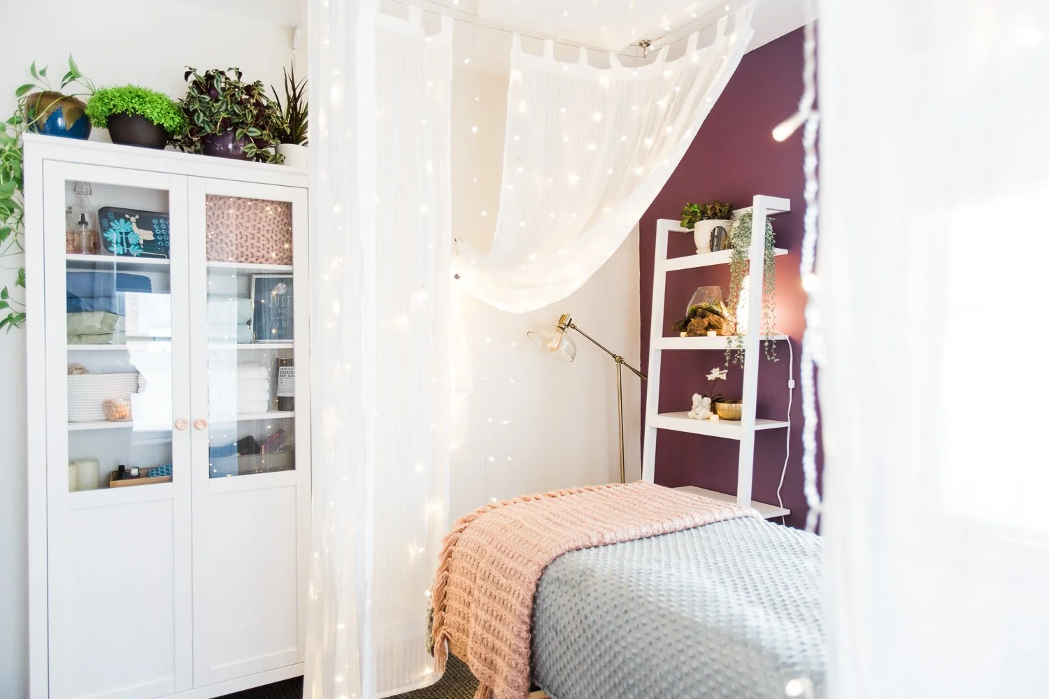 A cozy bedroom with a bed covered in a textured gray blanket and a pink throw blanket, a white bookshelf with plants and decorative items against a dark purple accent wall, a white storage cabinet with glass doors holding storage bins, and fairy ligh