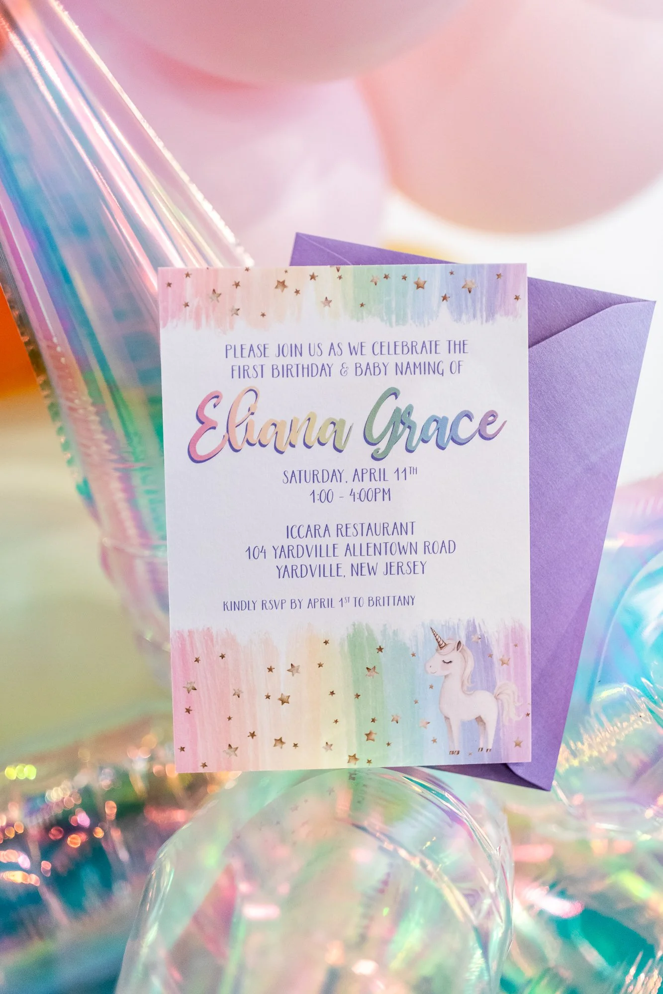 A colorful birthday invitation with pastel rainbow stripes, stars, and a cartoon unicorn, placed on an iridescent surface with floating, shiny balloons.