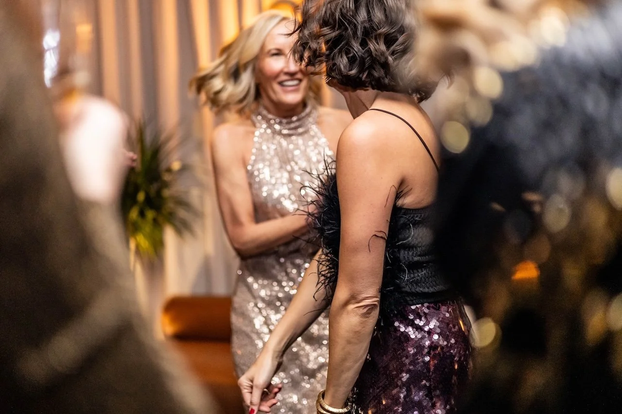 Two women in glamorous dresses are dancing and laughing at a party or celebration.